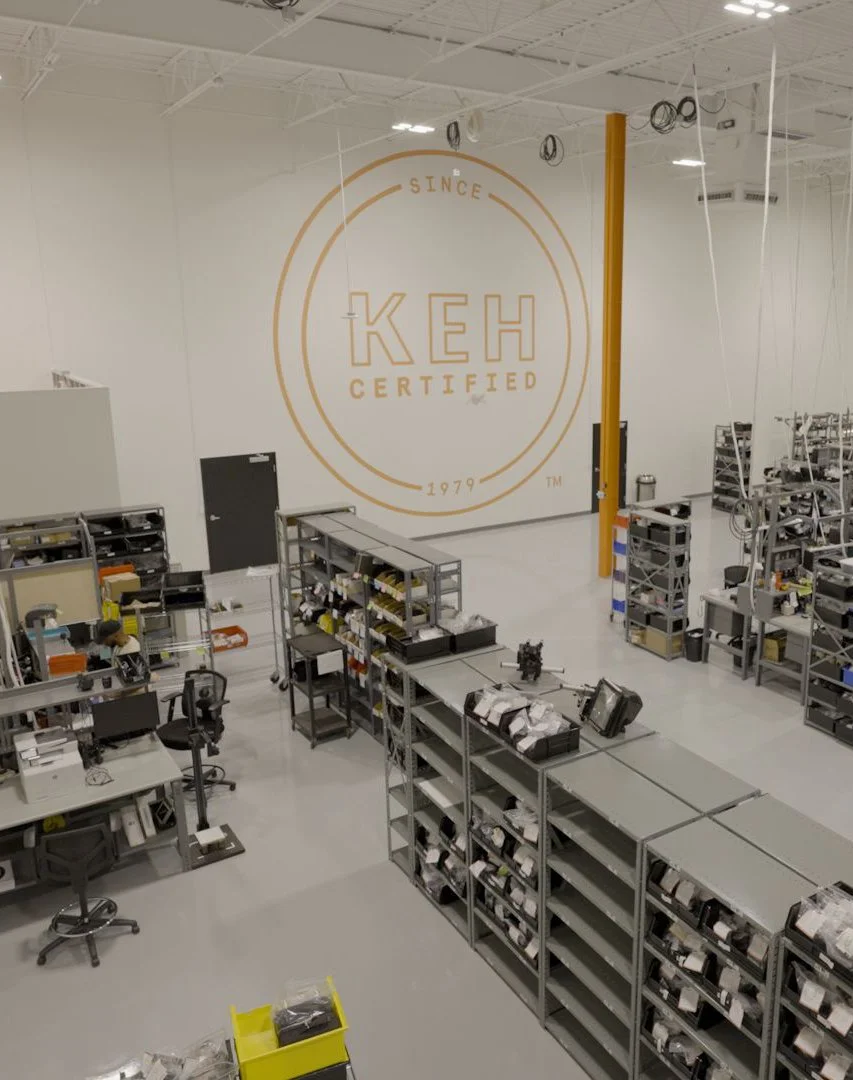 Manufacturing or warehouse space with metal shelves and workstations, large logo on the wall reading 'KEH Certified' with the words 'Since 1979'.