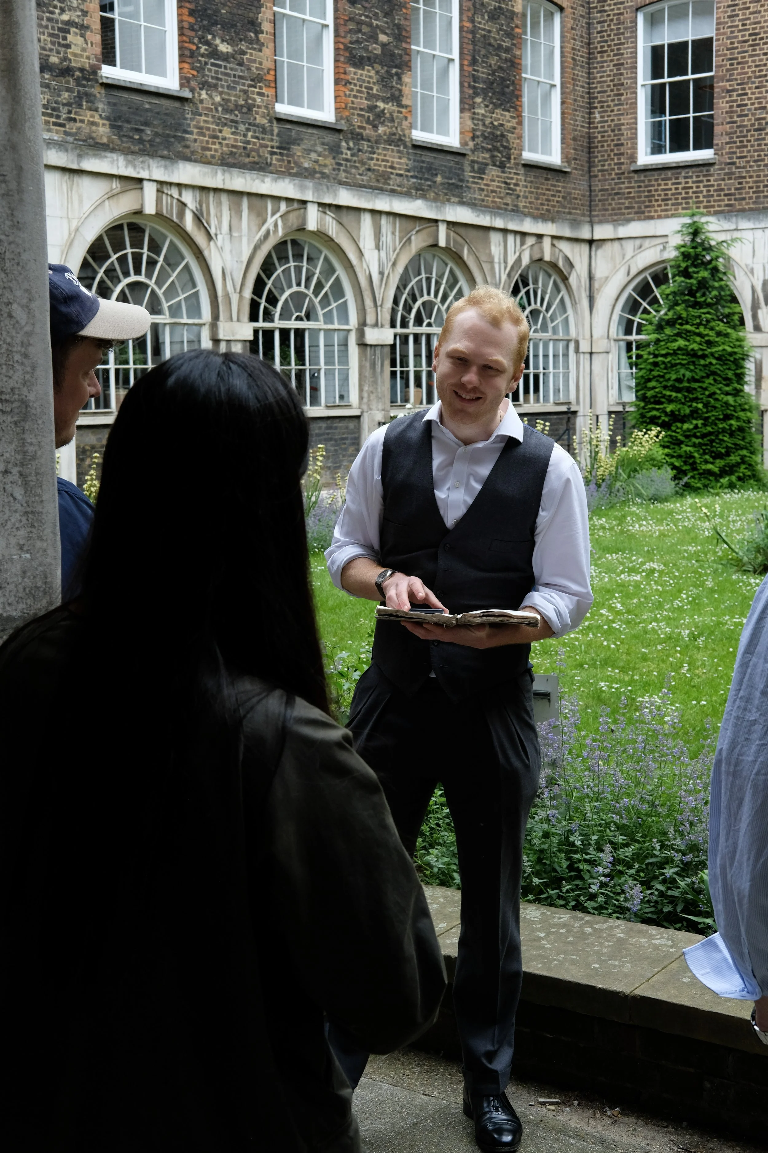 A man wearing a white shirt and black vest holding an open book, speaking to a small group of people outdoors in a garden area with a brick building with arched windows in the background.