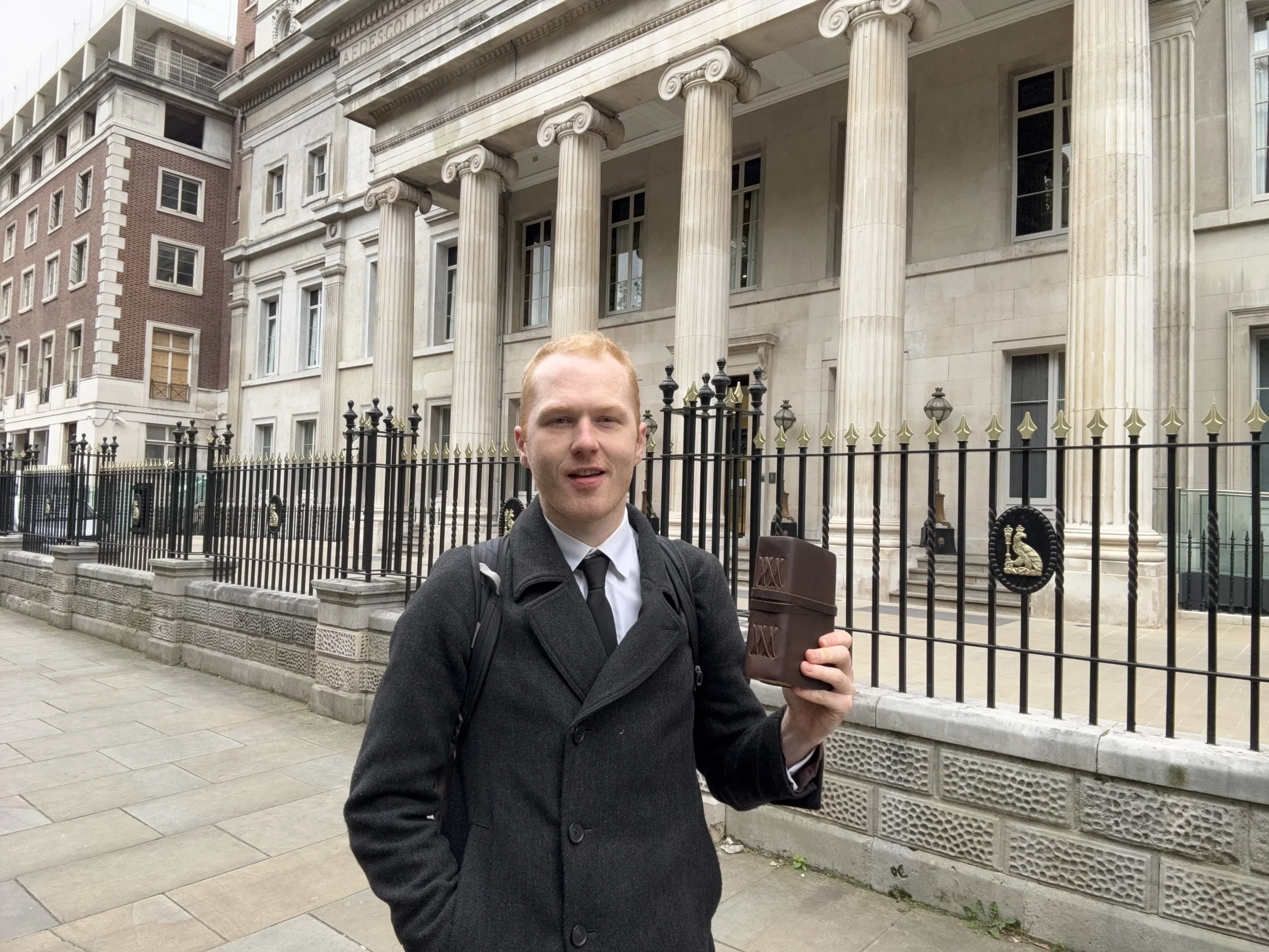 A man with light skin and red hair wearing a black coat and carrying a backpack stands on a city sidewalk, holding a small brown leather-bound notebook or planner, in front of a historic building with large columns and a black iron fence.