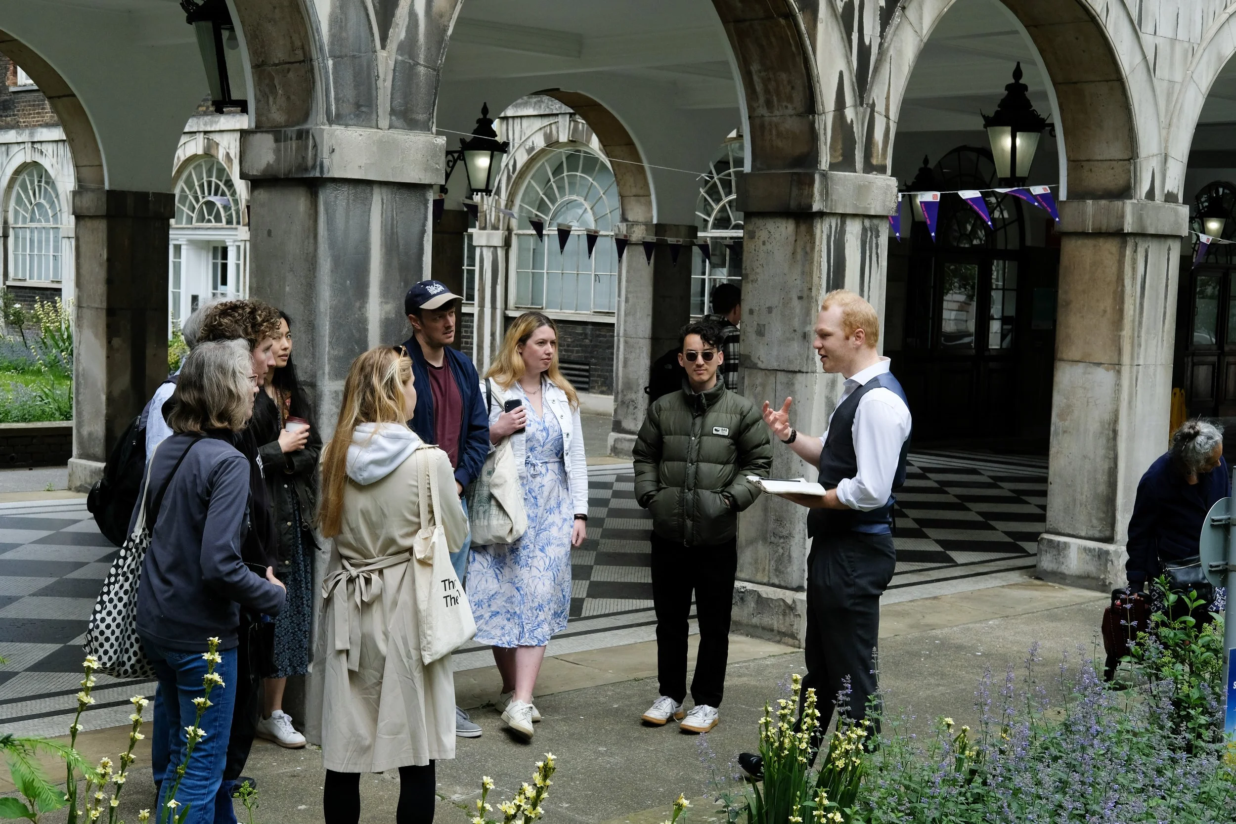 A tour guide speaking to a group of diverse tourists standing under archways on a city street, with flowers in the foreground and historical buildings in the background.