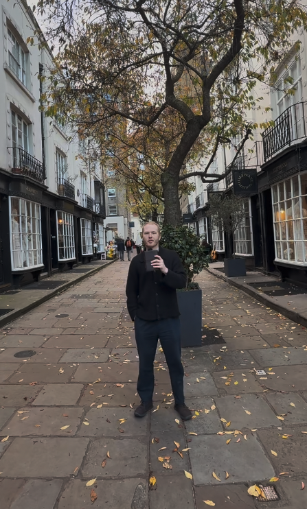 Person taking a selfie on a city street lined with buildings and a large tree in a planter.