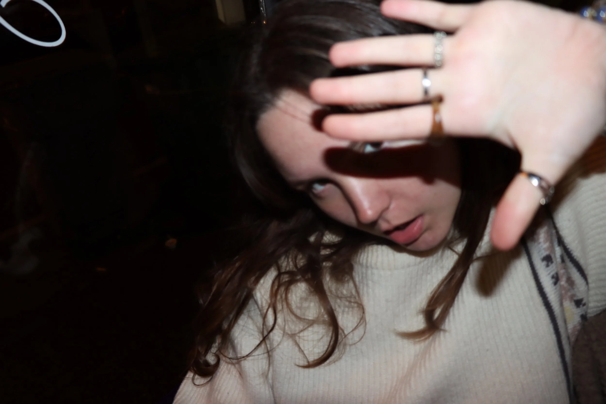 A woman with long brown hair covering part of her face with her left hand, wearing multiple rings, in a dimly lit environment.