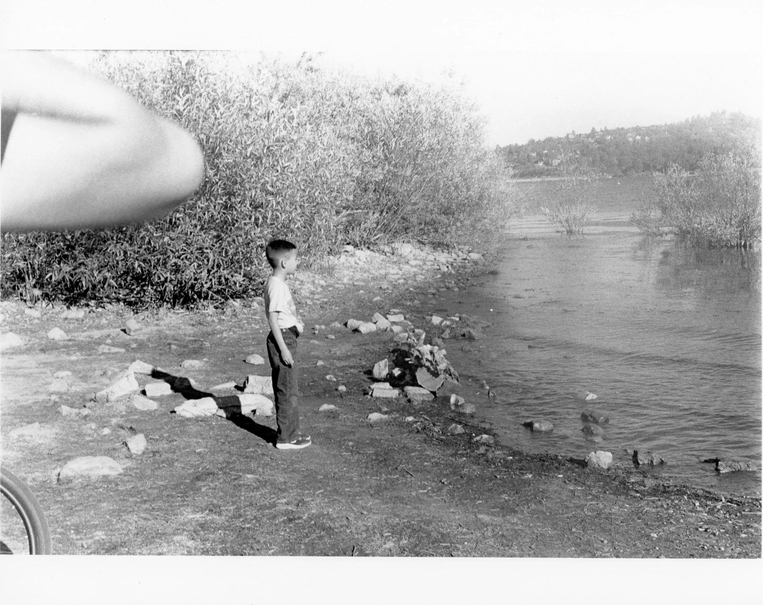 A young boy standing on a rocky lakeshore, looking out at the water with hills and trees in the background, black and white photo.