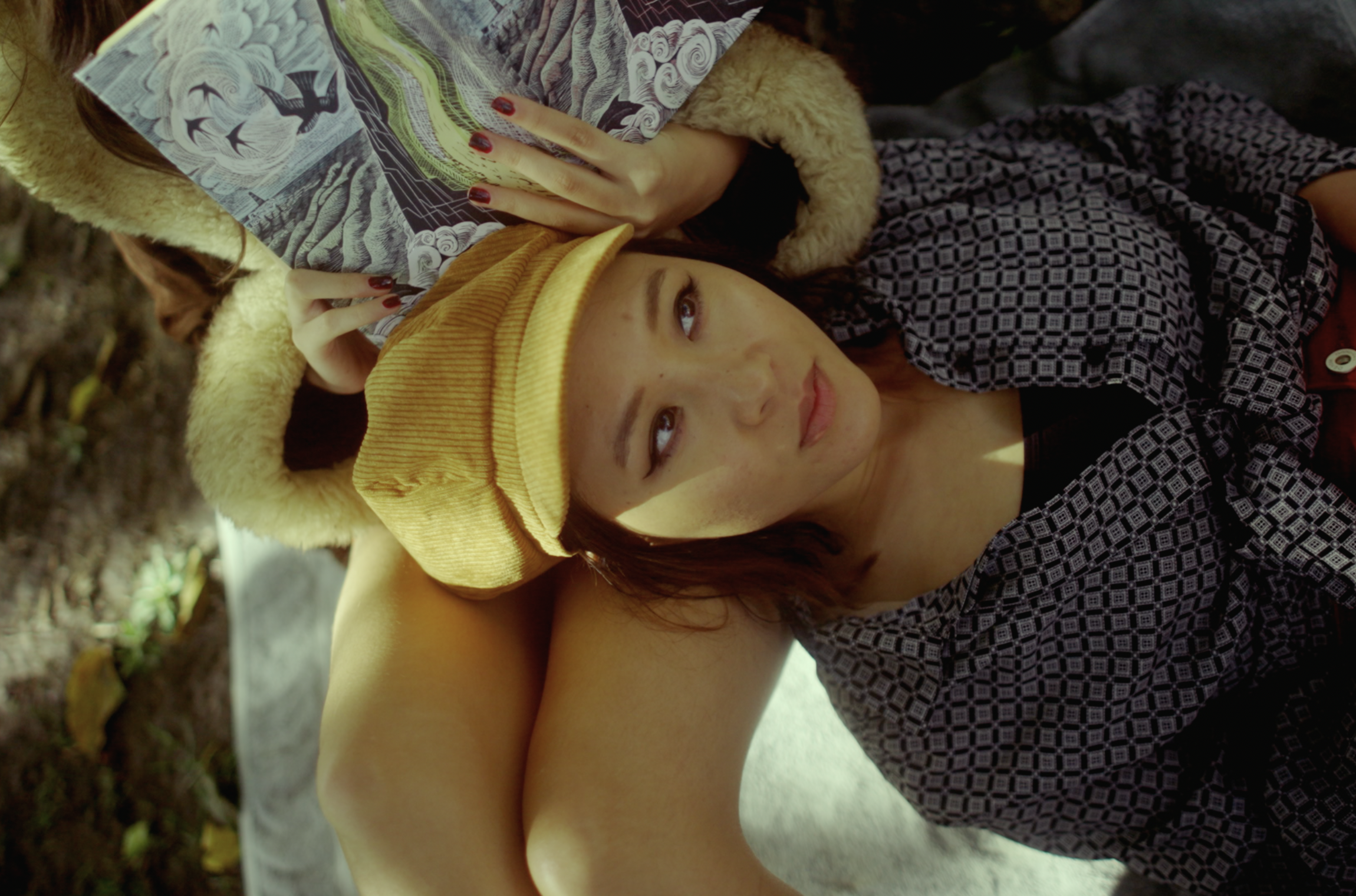 A young woman with red hair and light skin, lying outdoors on a stone surface, wearing a yellow beret and a patterned shirt, holding a book or magazine and looking up.