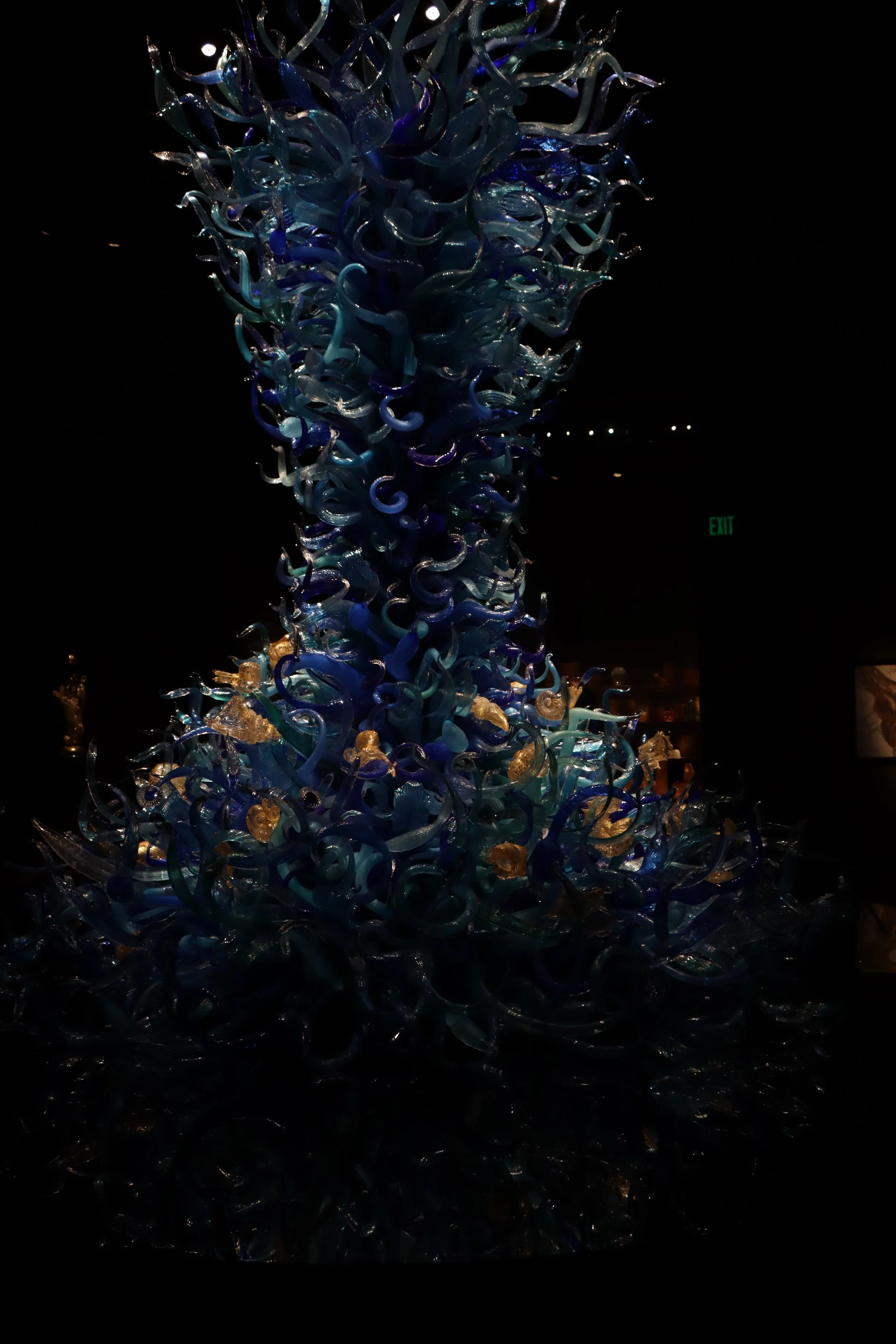 A large, intricate art sculpture made of blue and clear glass, shaped into swirling, twisted forms, with some gold-colored glass accents at the base, displayed in a dark exhibition space.