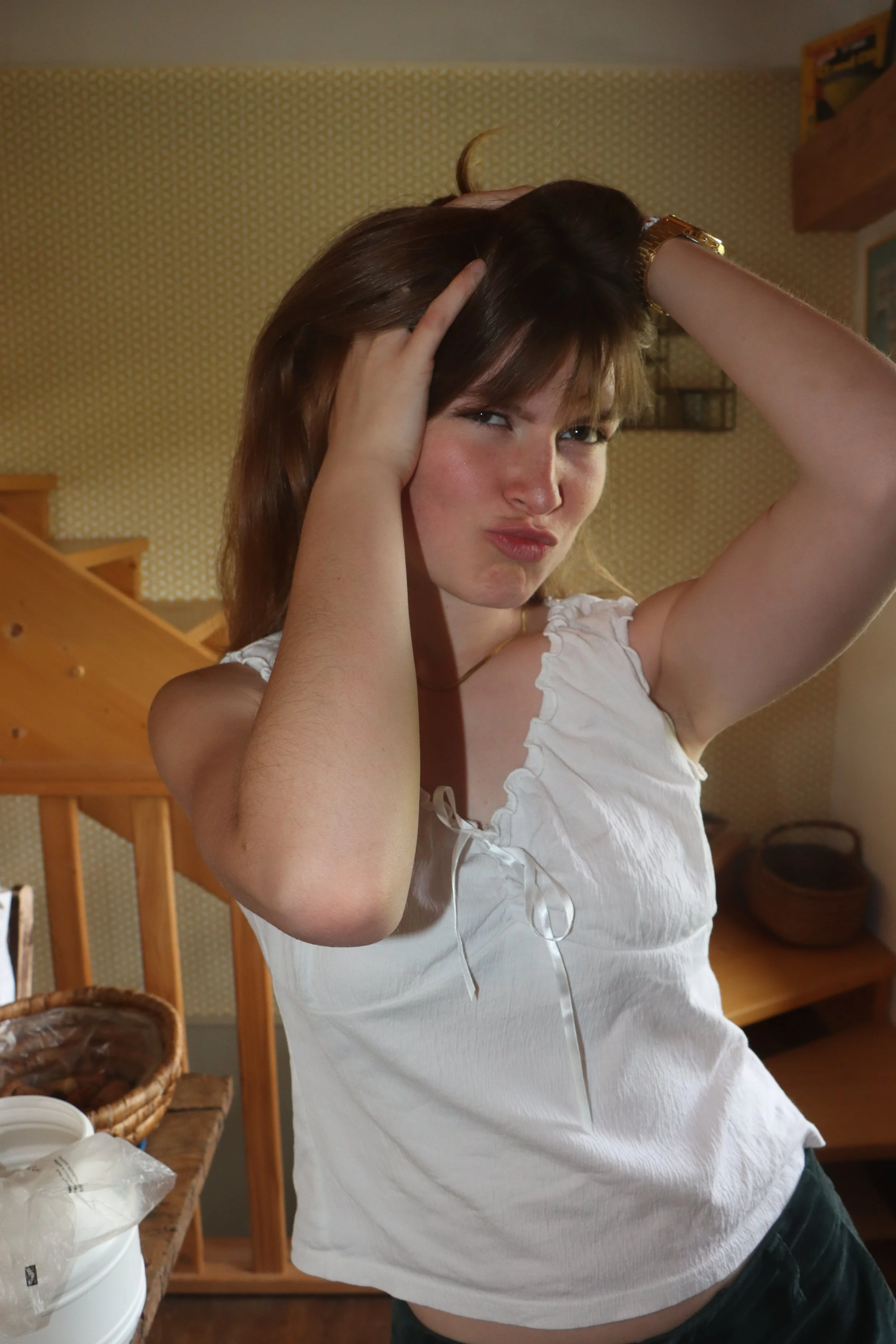 A woman with brown hair wearing a white sleeveless top is standing in a room with wooden stairs and furniture. She has her hands in her hair and is making a playful, pouting facial expression.