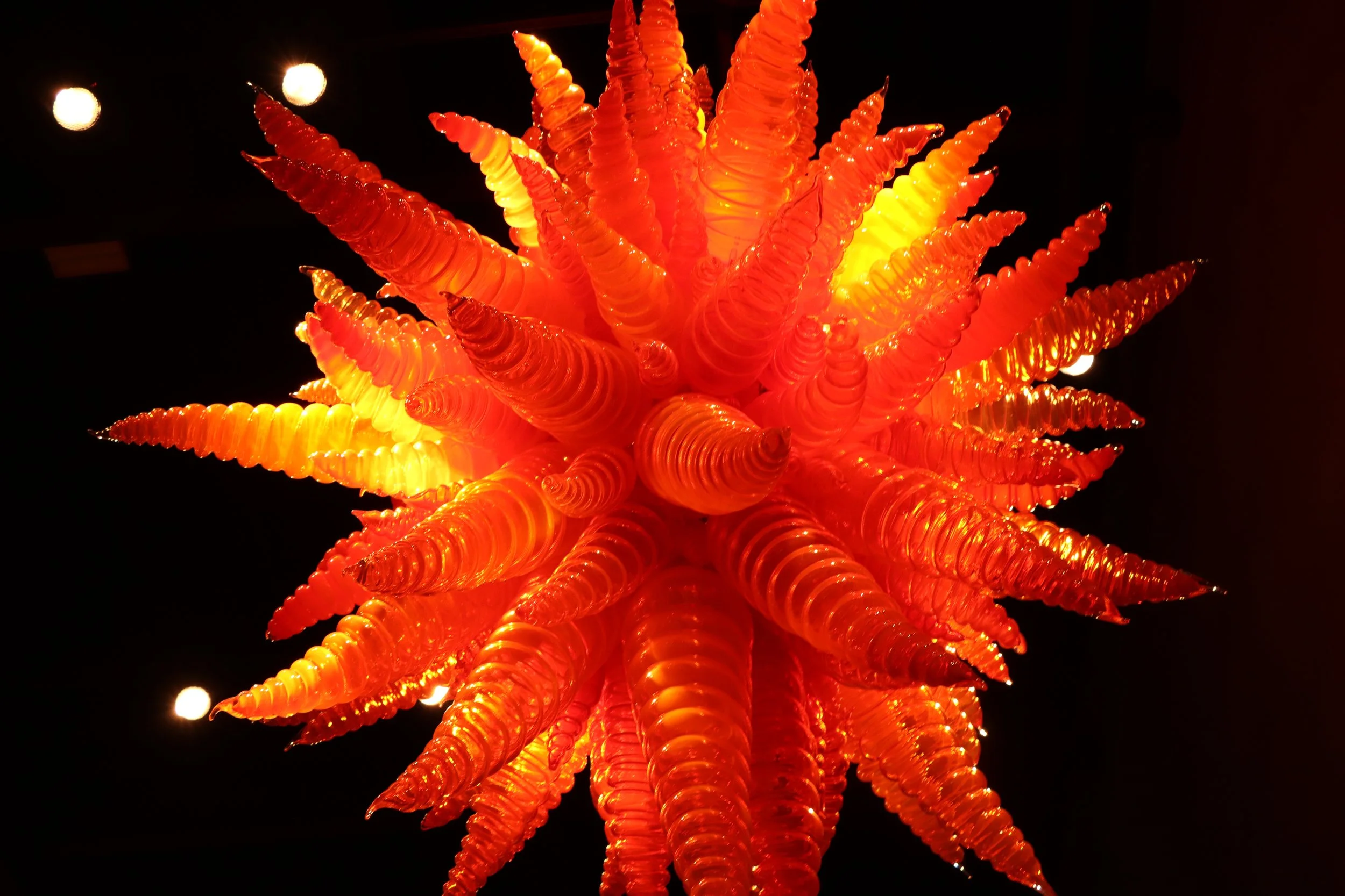 Colorful glass art sculpture resembling a starburst with orange and yellow spiraled cones against a black background.
