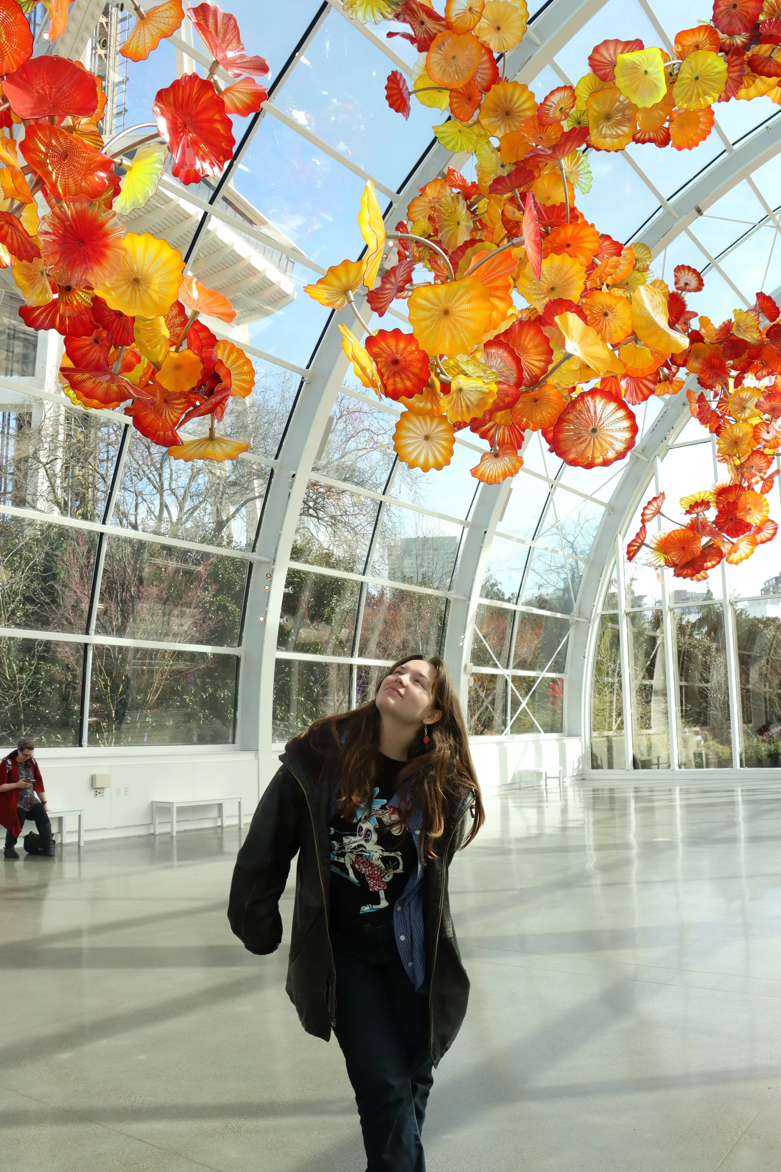 A woman stands inside a glass art installation with her eyes closed, looking up at a large, colorful glass sculpture of orange, red, and yellow flowers or leaves hanging overhead. The background shows trees and a blue sky visible through the glass ceiling.