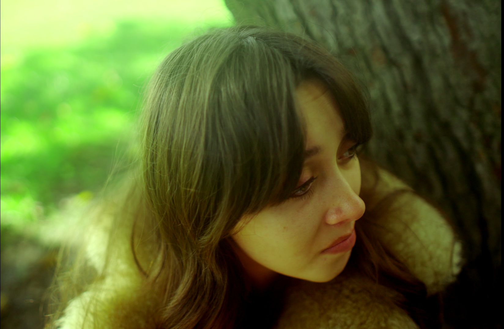Close-up of a young woman with long, dark hair, leaning against a tree with a blurred green background.
