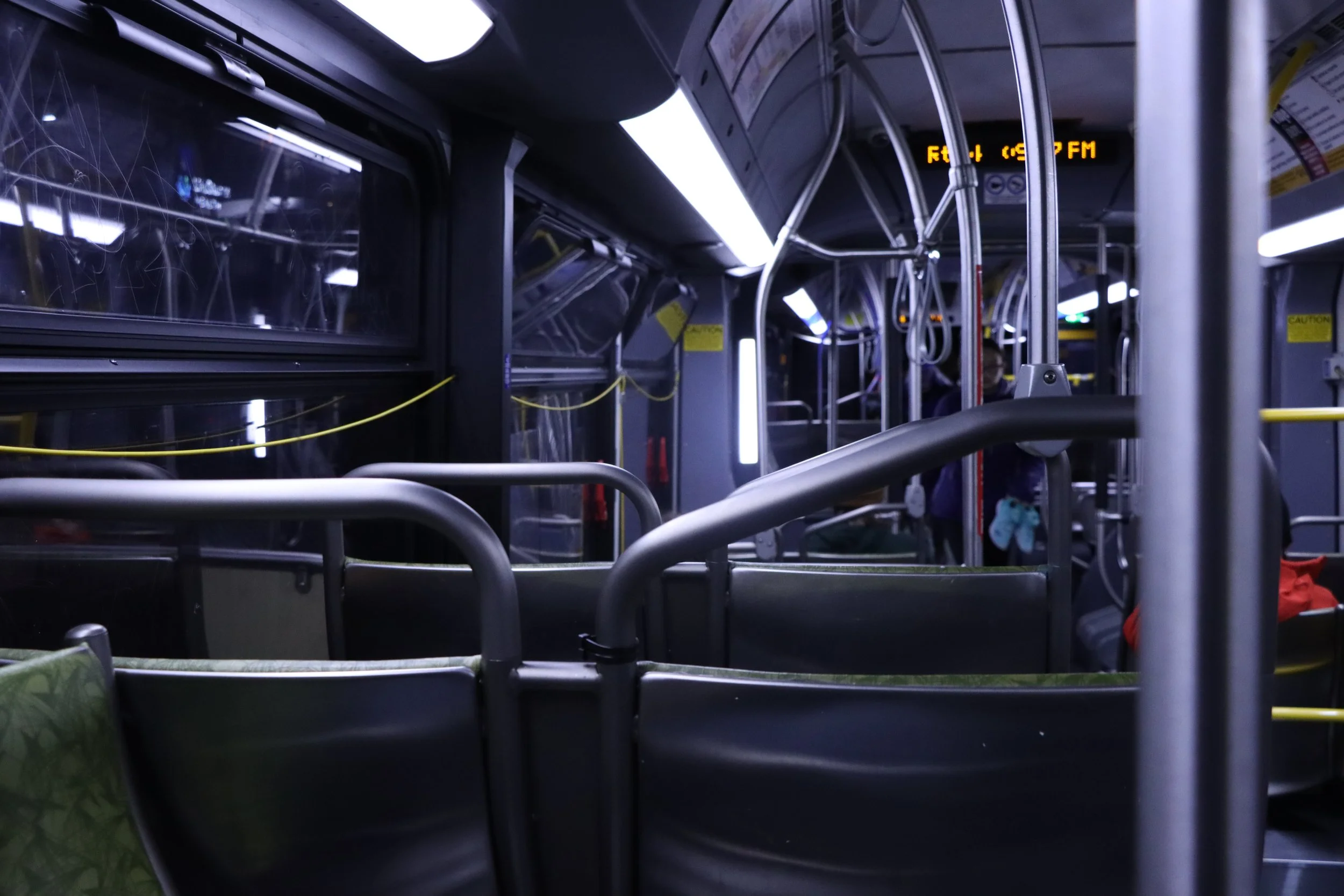 Inside a city bus with empty seats and metal handrails, illuminated by interior lighting, with some passengers in the background.