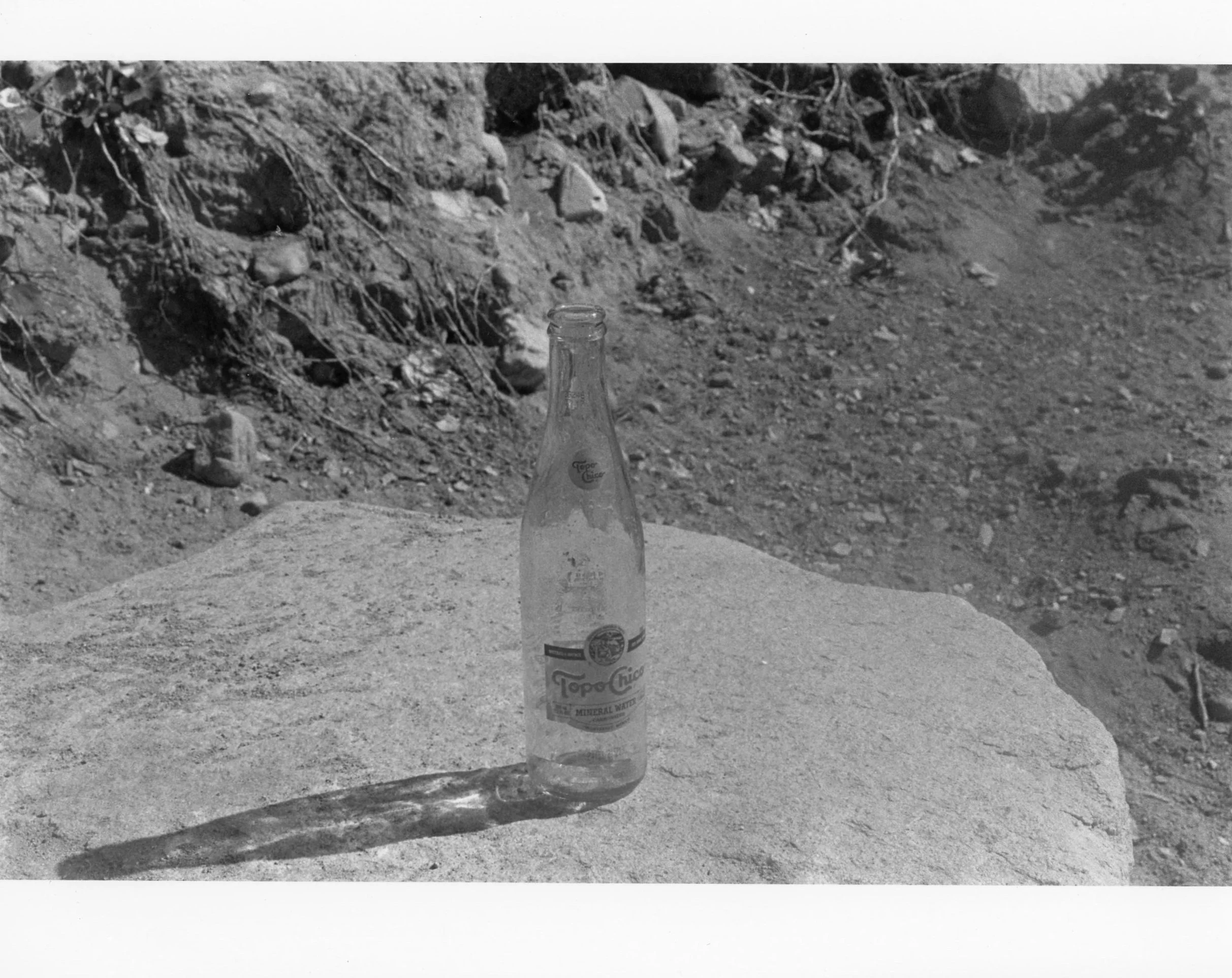 A clear glass Topo Chico mineral water bottle placed on a flat rock outdoors, casting a long shadow on the rock surface.