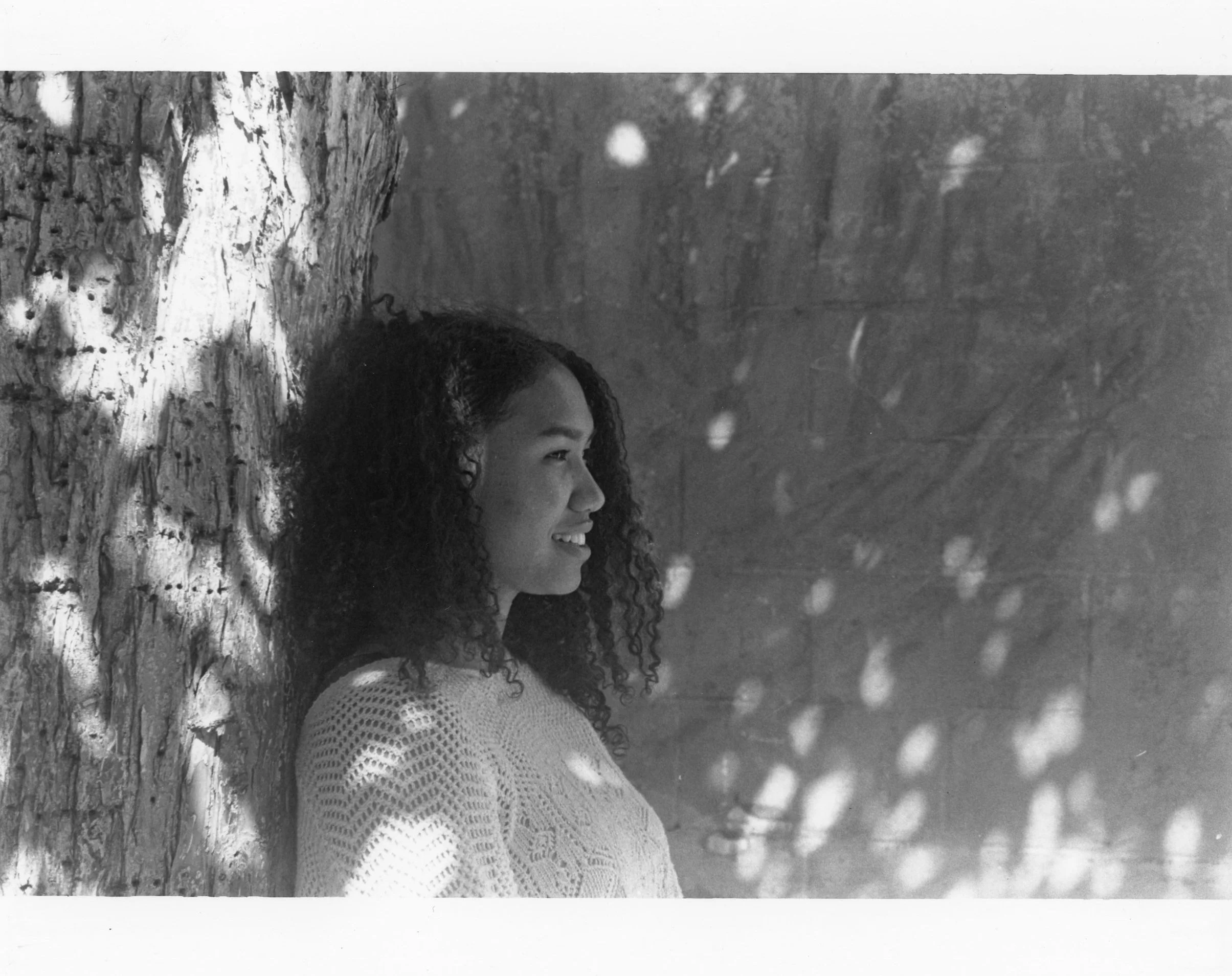 Black and white photo of a young woman with curly hair smiling and leaning against a tree trunk, with sunlight creating dappled shadows on her face and background.