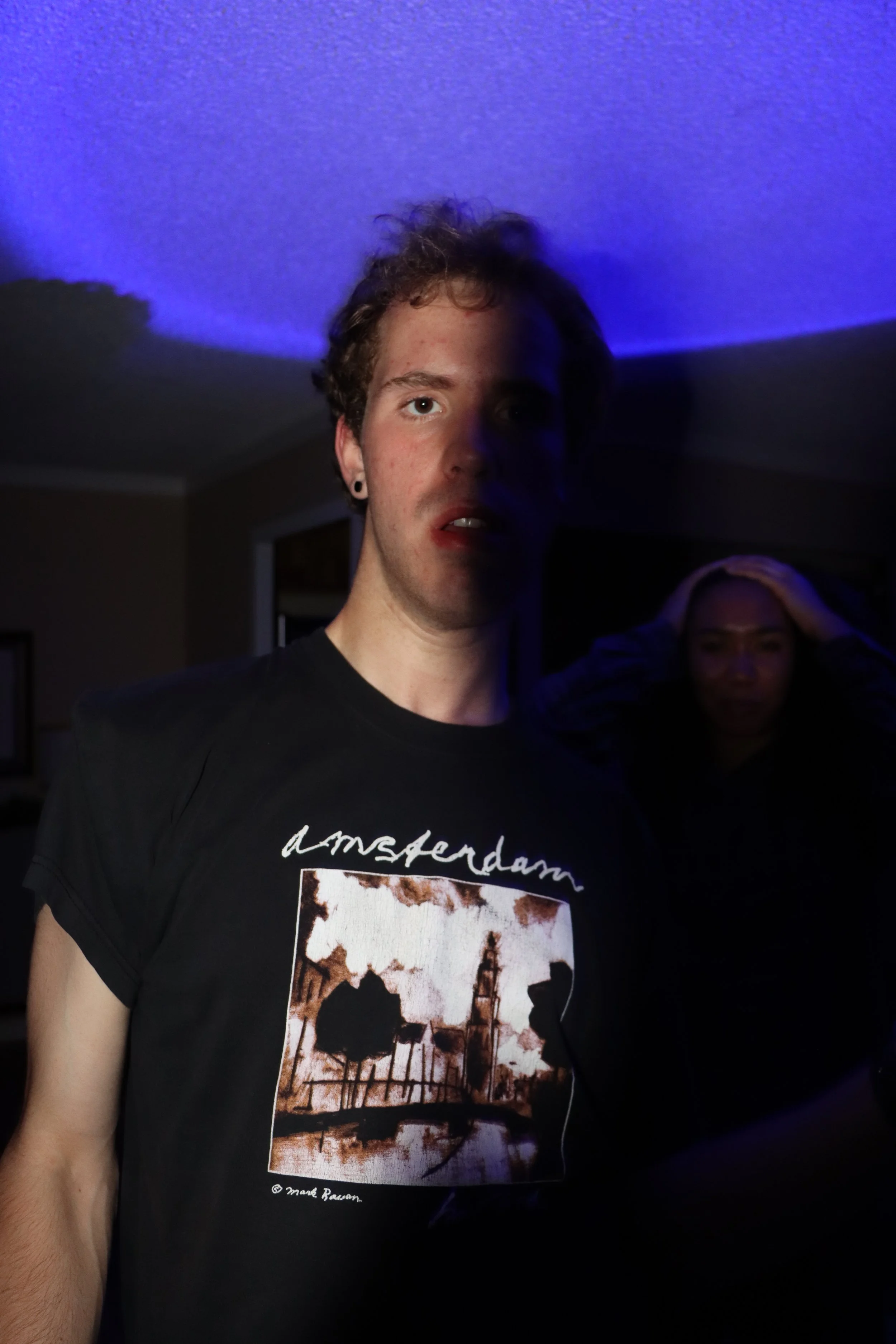 A young man with curly hair and earrings taking a selfie in a dimly lit room with purple and blue lighting, with a woman in the background holding her head.