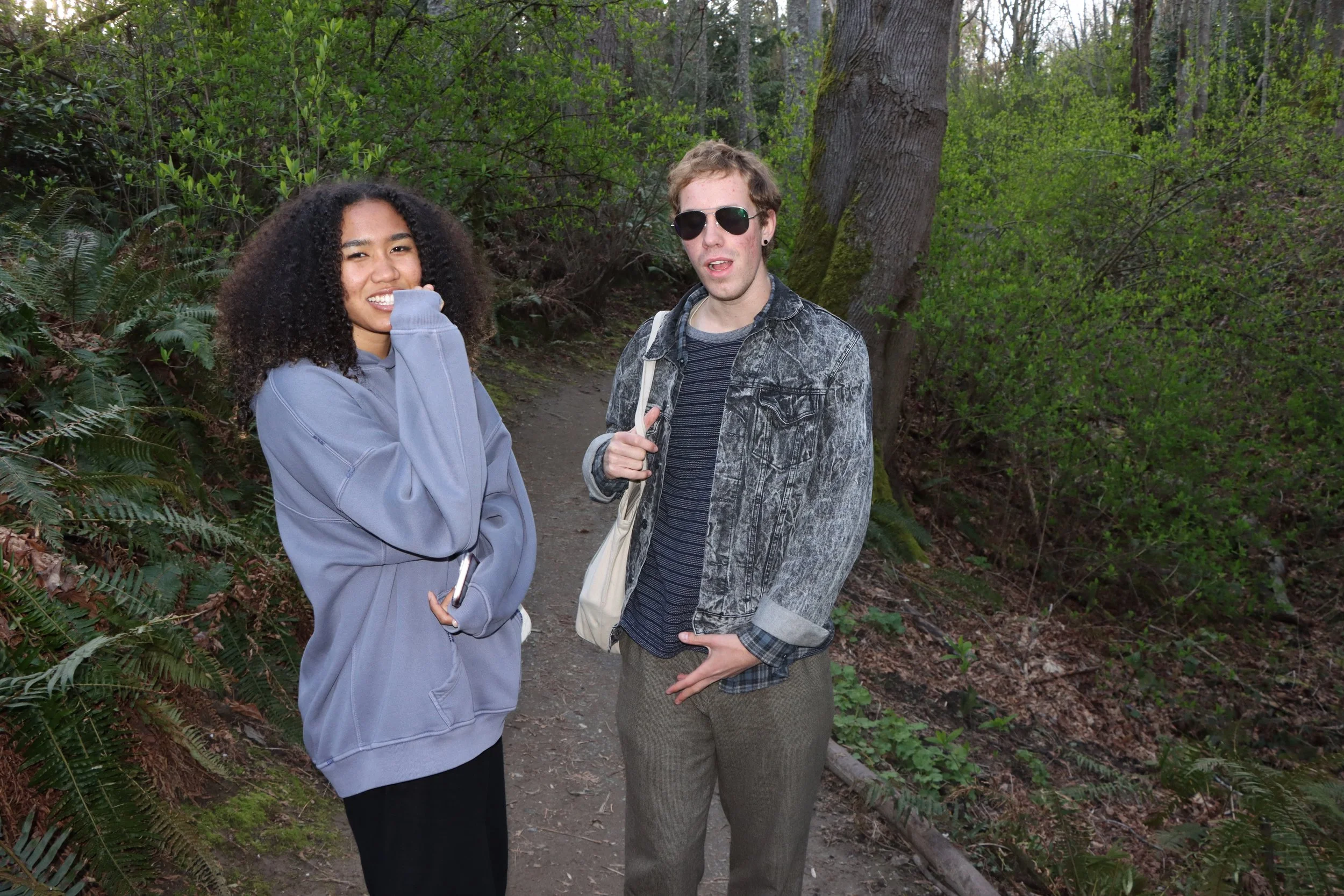 A young woman with curly hair smiling and wearing a gray hoodie, standing next to a young man with sunglasses, short hair, and a gray denim jacket, on a forest trail surrounded by green trees and foliage.