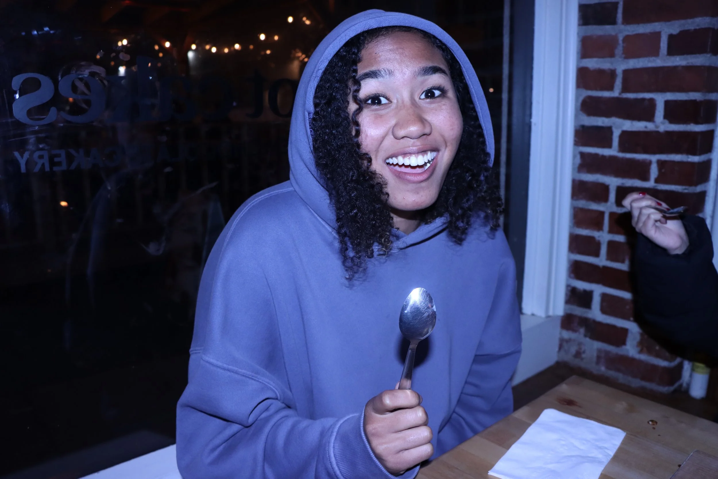 Young woman with curly hair smiling and holding a spoon, sitting at a table in a restaurant or cafe at night.