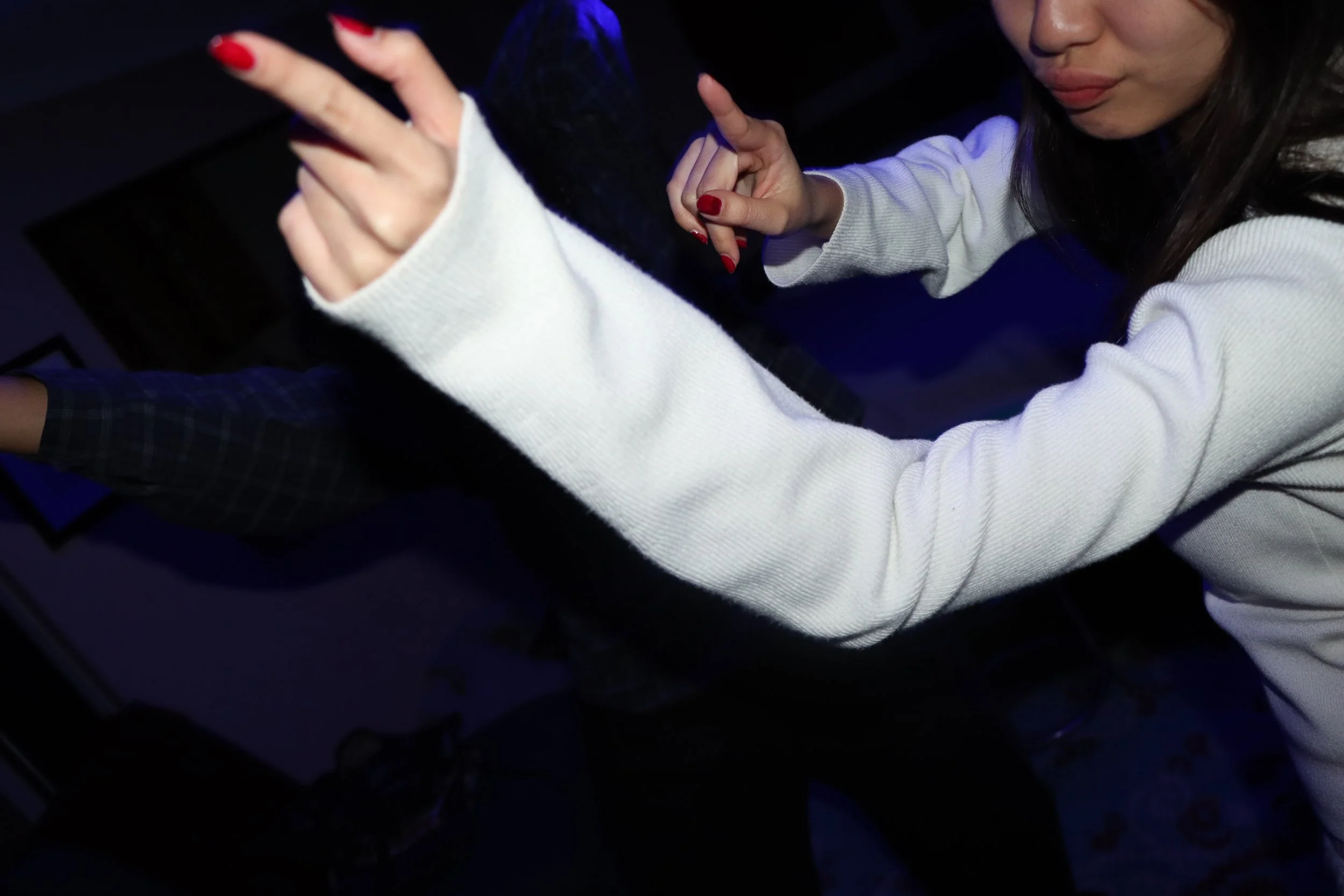 A woman with dark hair taking a selfie in a dark room, wearing a white sweater with red nail polish, and showing a peace sign with her hand.