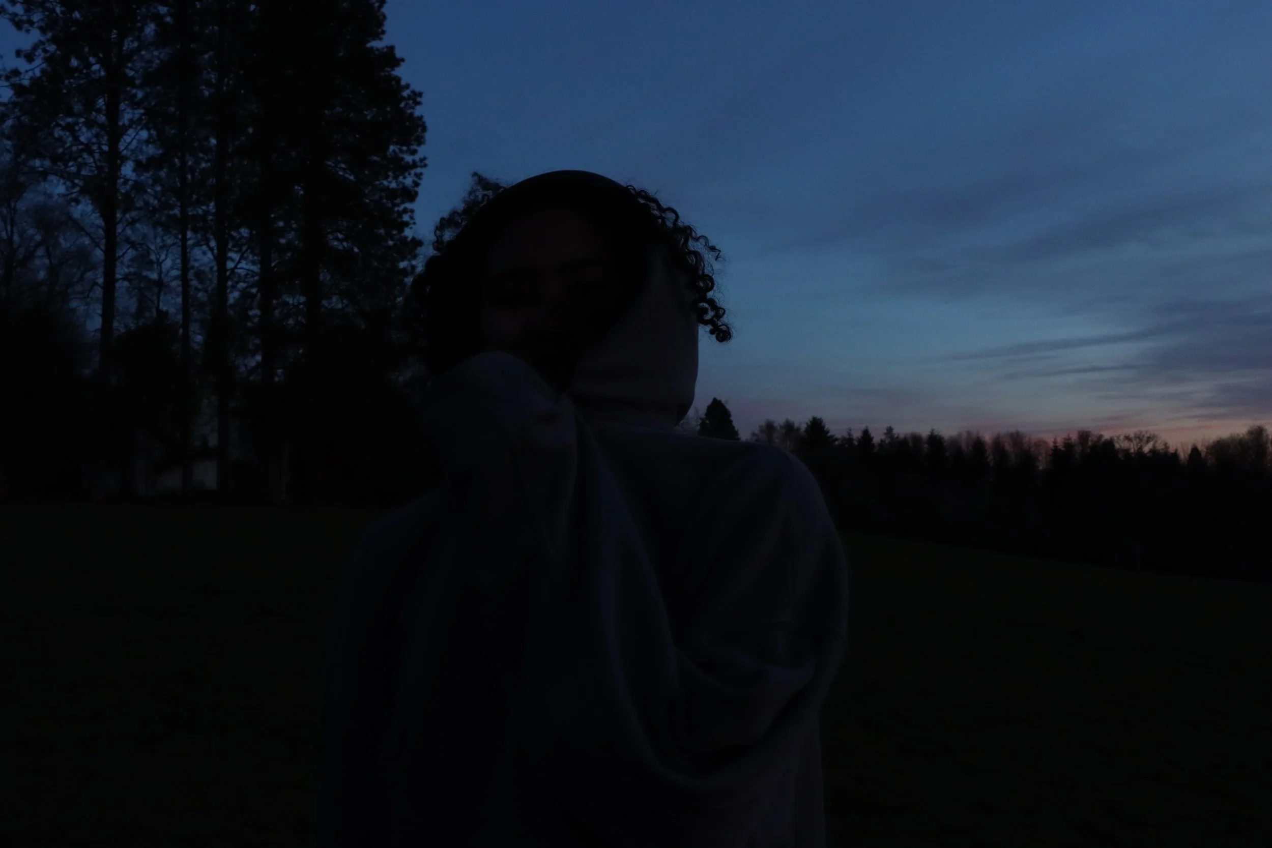 A person standing outdoors during sunset or dusk with a sky that has colorful streaks and silhouette trees in the background.