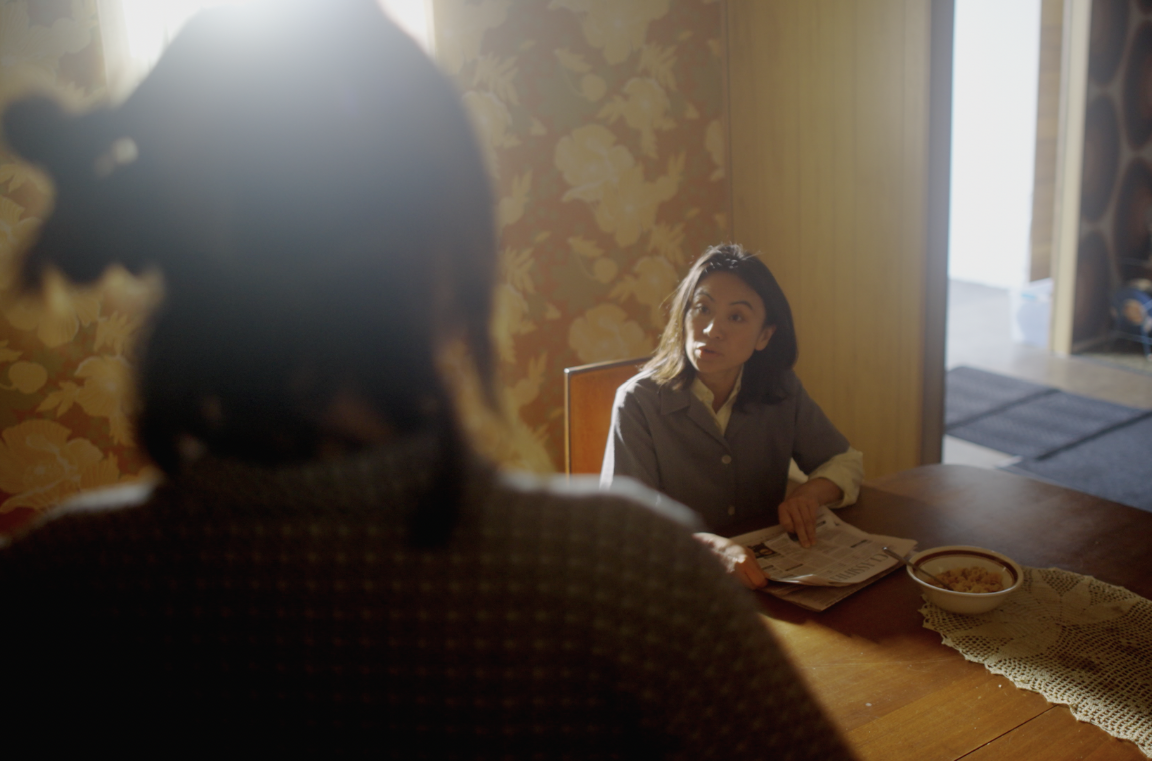 A woman sitting at a dining table with a bowl of cereal, talking to another person whose back is to the camera, in a sunlit room with floral wallpaper and a doorway leading outside.