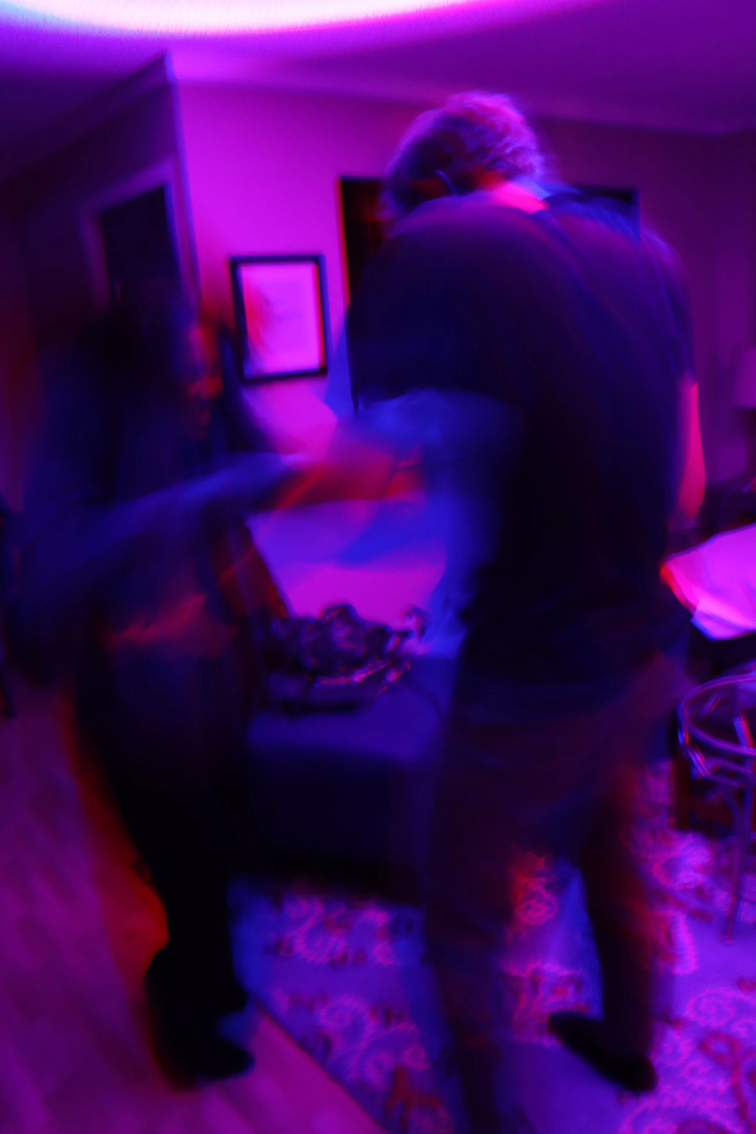 A blurry photo of two people dancing or socializing in a room illuminated with purple and pink neon lights.