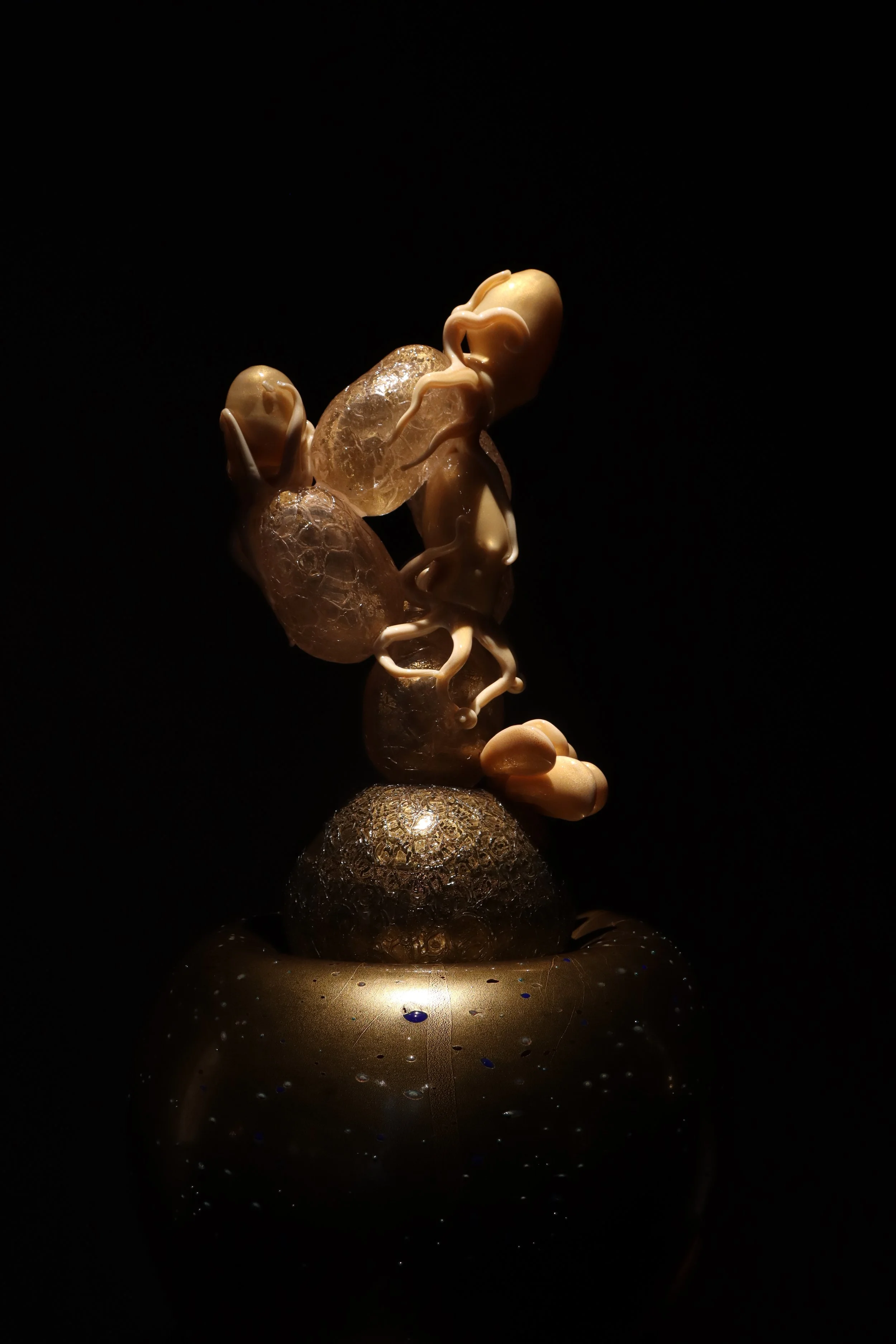 A sculpture featuring abstract shapes and organic forms illuminated against a dark background, composed of smooth, rounded elements and textured surfaces with a reflective base.