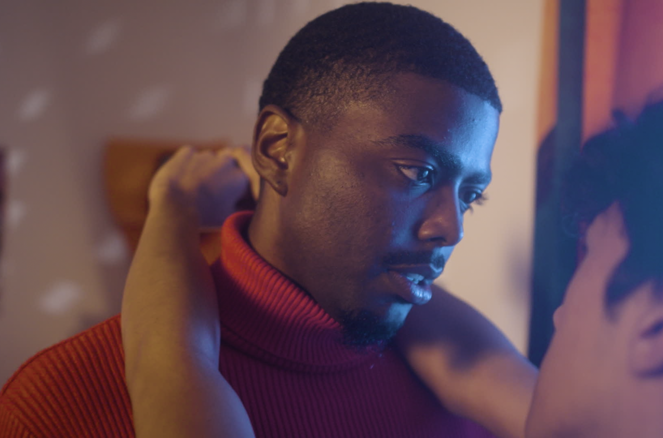A young man with short hair and a beard looking down, wearing a red turtleneck sweater, with someone behind him touching his neck. The background has warm lighting and soft focus.