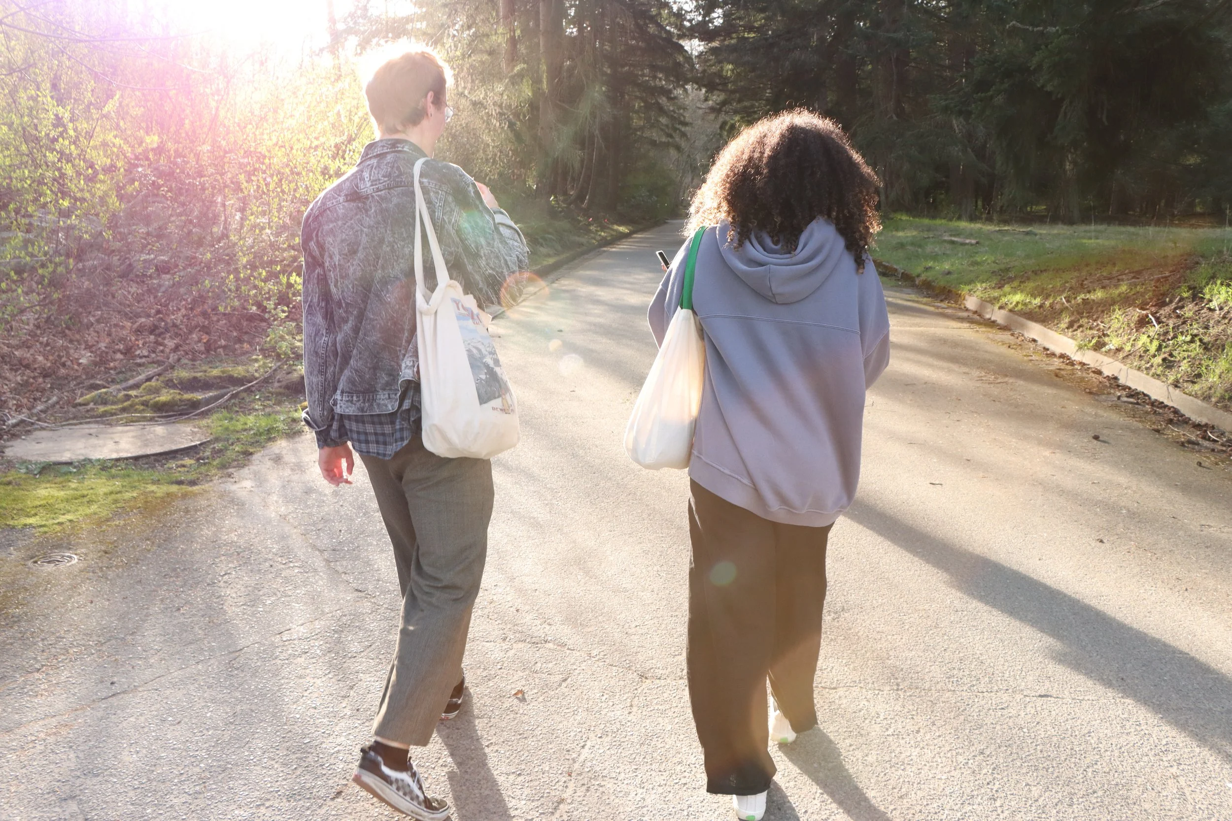 Two young women walking on a sunlit forest path, one wearing a gray hoodie and the other wearing a denim jacket, both carrying tote bags.