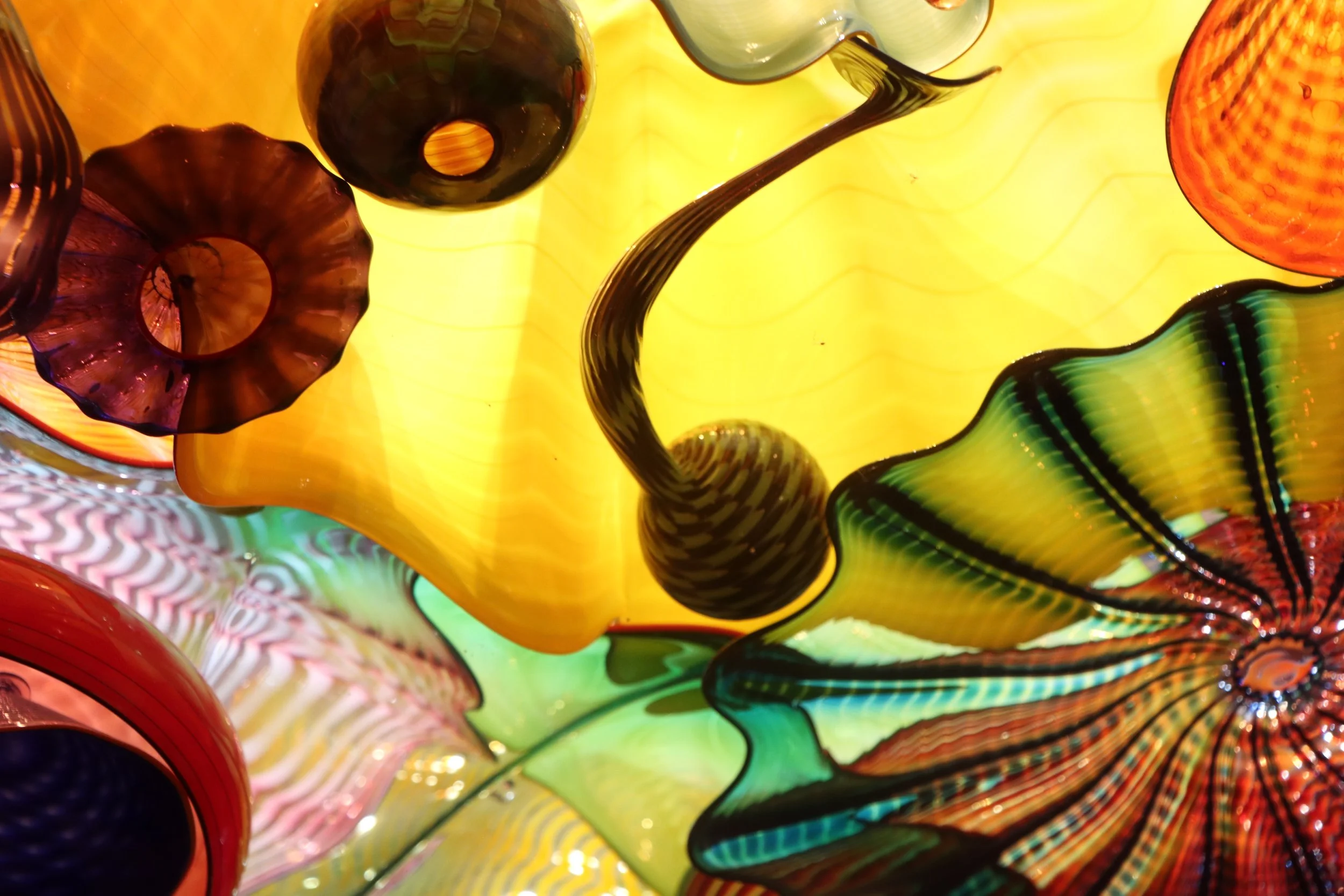 Colorful blown glass art with various shapes and patterns, illuminated with a yellow background.