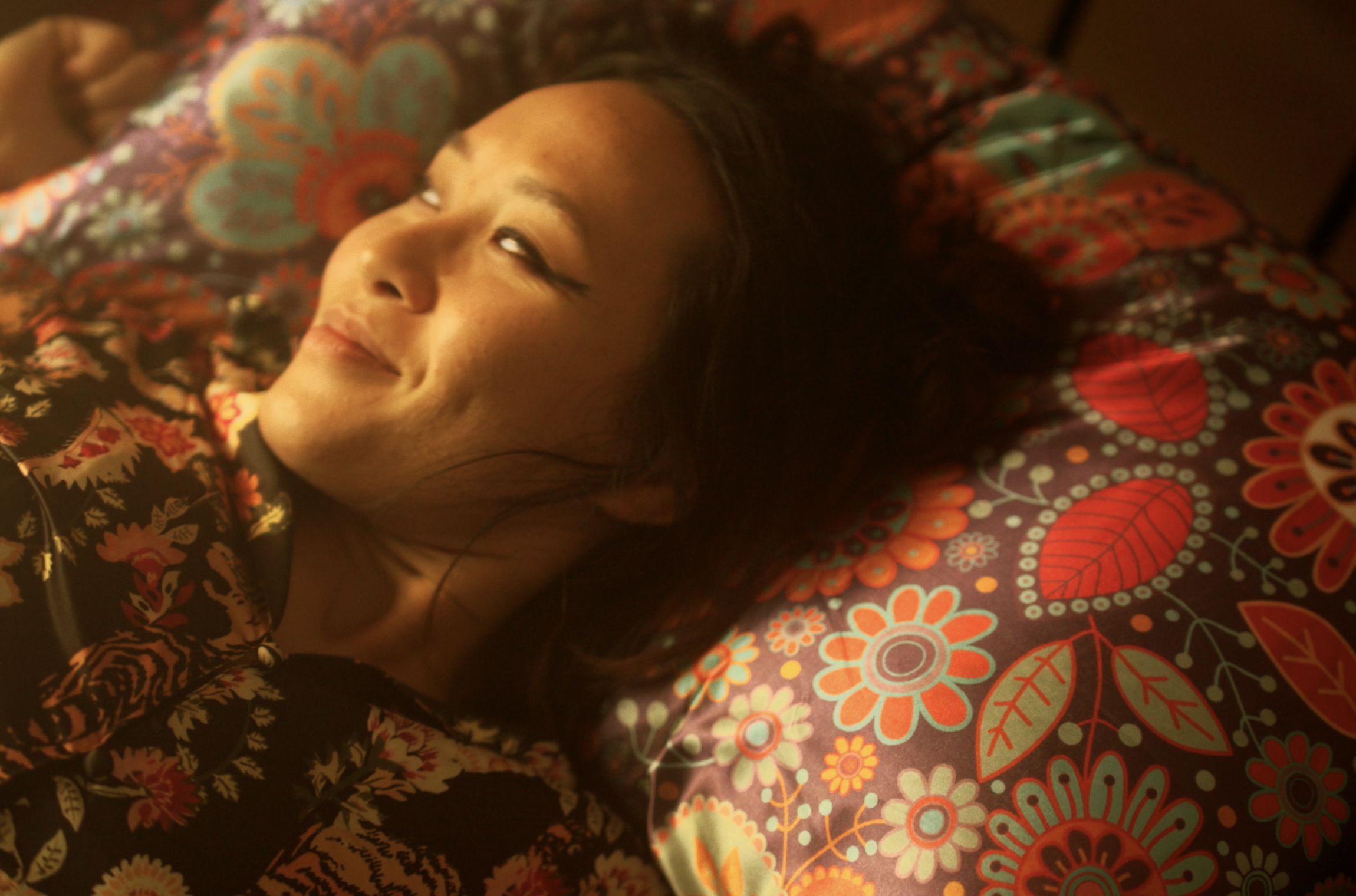 A woman with a gentle smile lying on a colorful floral pillow, wearing a patterned top.