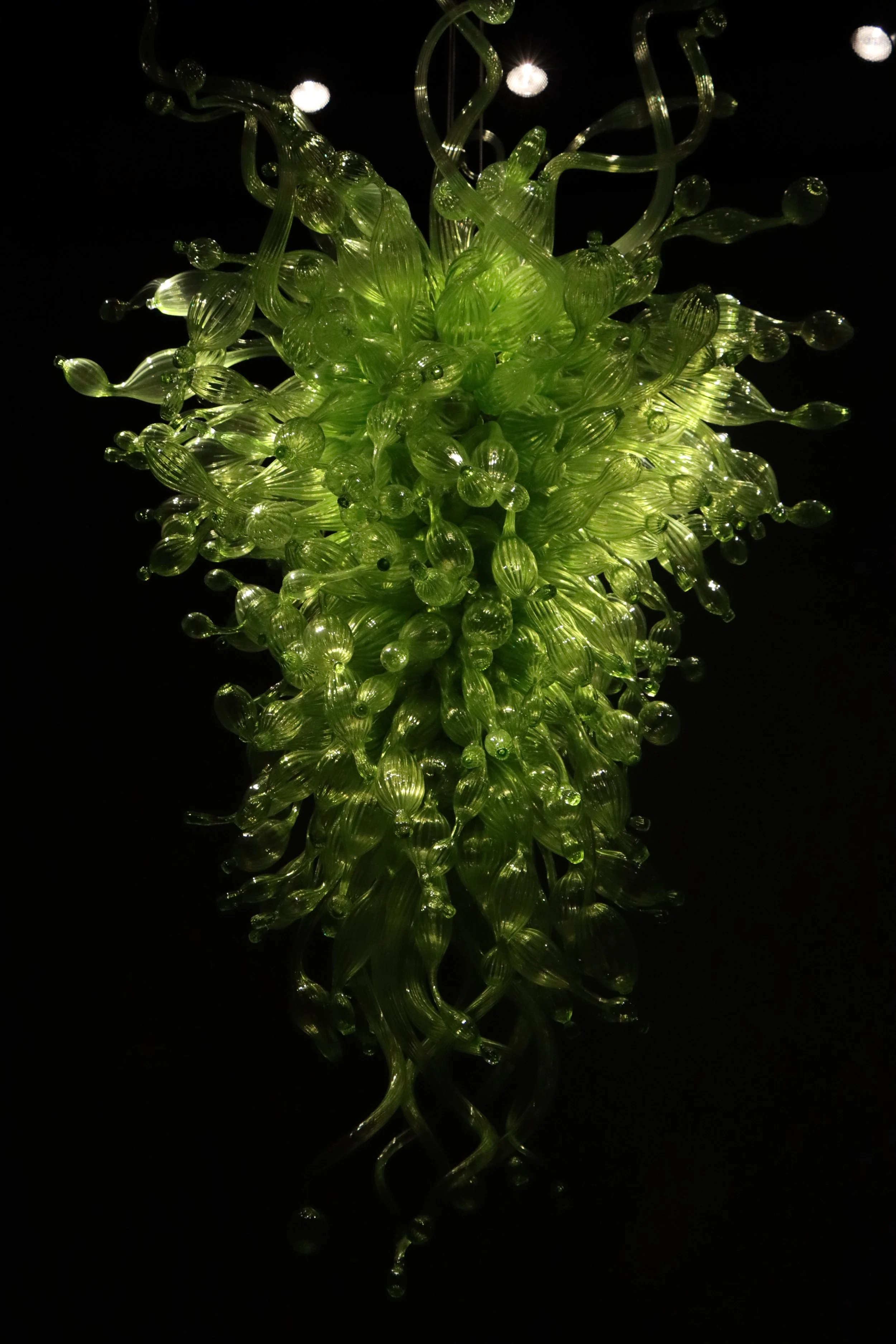 A large, intricate green glass chandelier with twisting, elongated glass pieces, hanging against a dark background.
