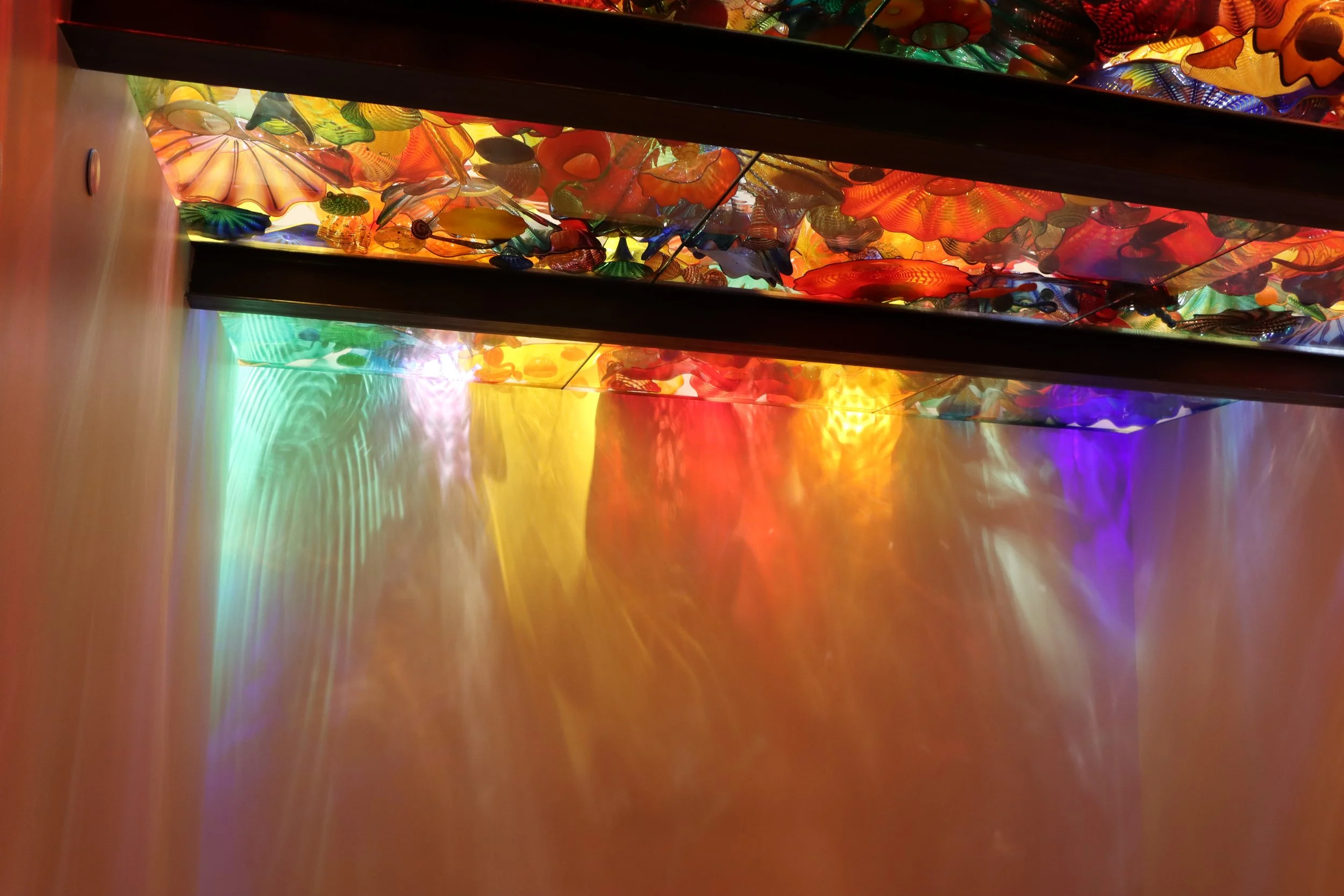 Colorful glass art installation mounted on the ceiling, casting multi-colored reflections onto a shiny surface below.