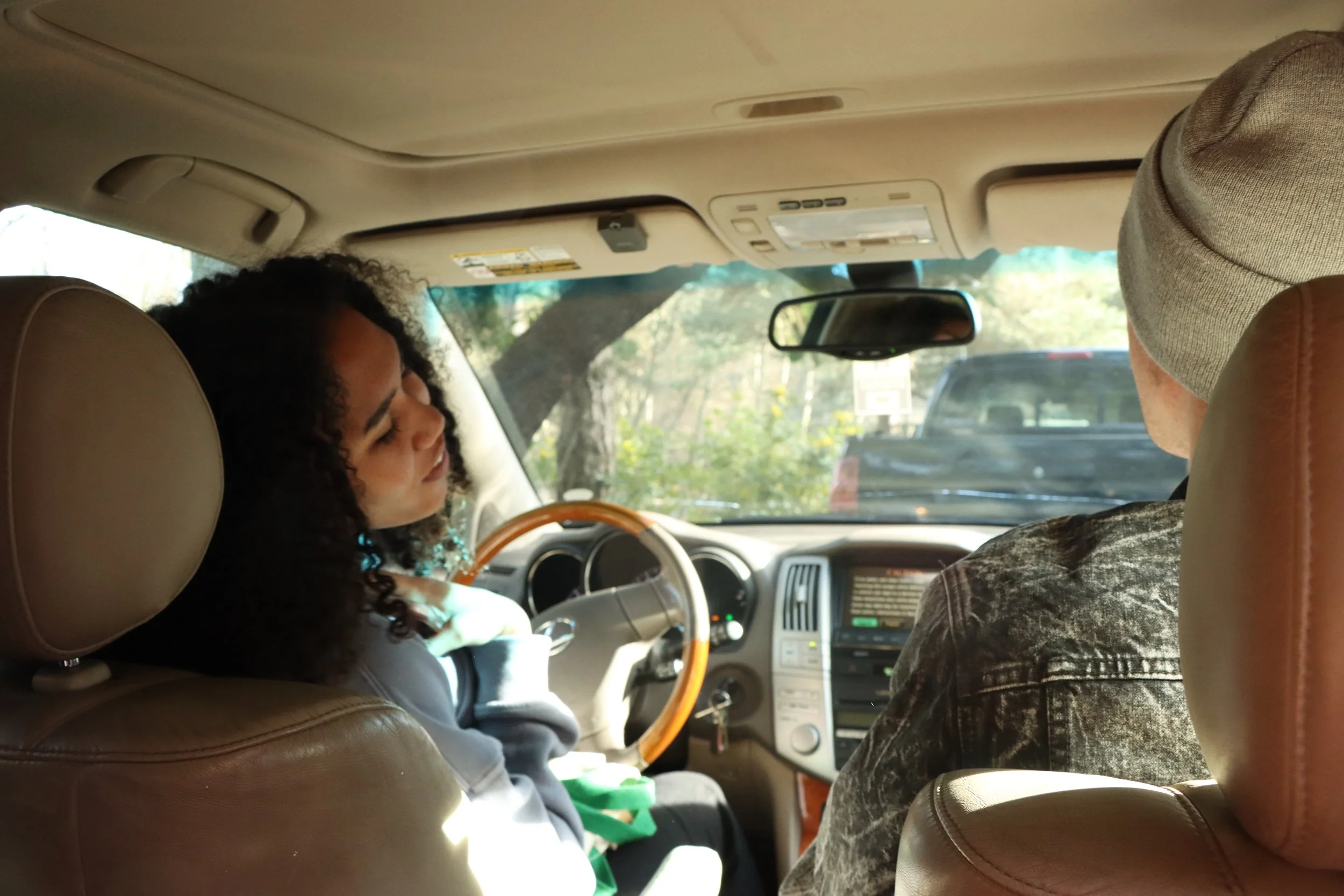 Two people sitting in the front seats of a vehicle, with the woman on the driver’s side leaning back and the man on the passenger’s side facing forward, both facing towards the front of the vehicle. The vehicle's interior includes the dashboard, steering wheel, and rearview mirror, with trees visible outside through the windshield.