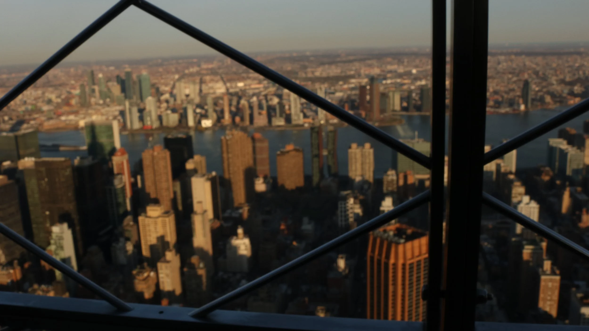 A city skyline viewed from a high vantage point, with skyscrapers and a river visible, framed by metal scaffolding.