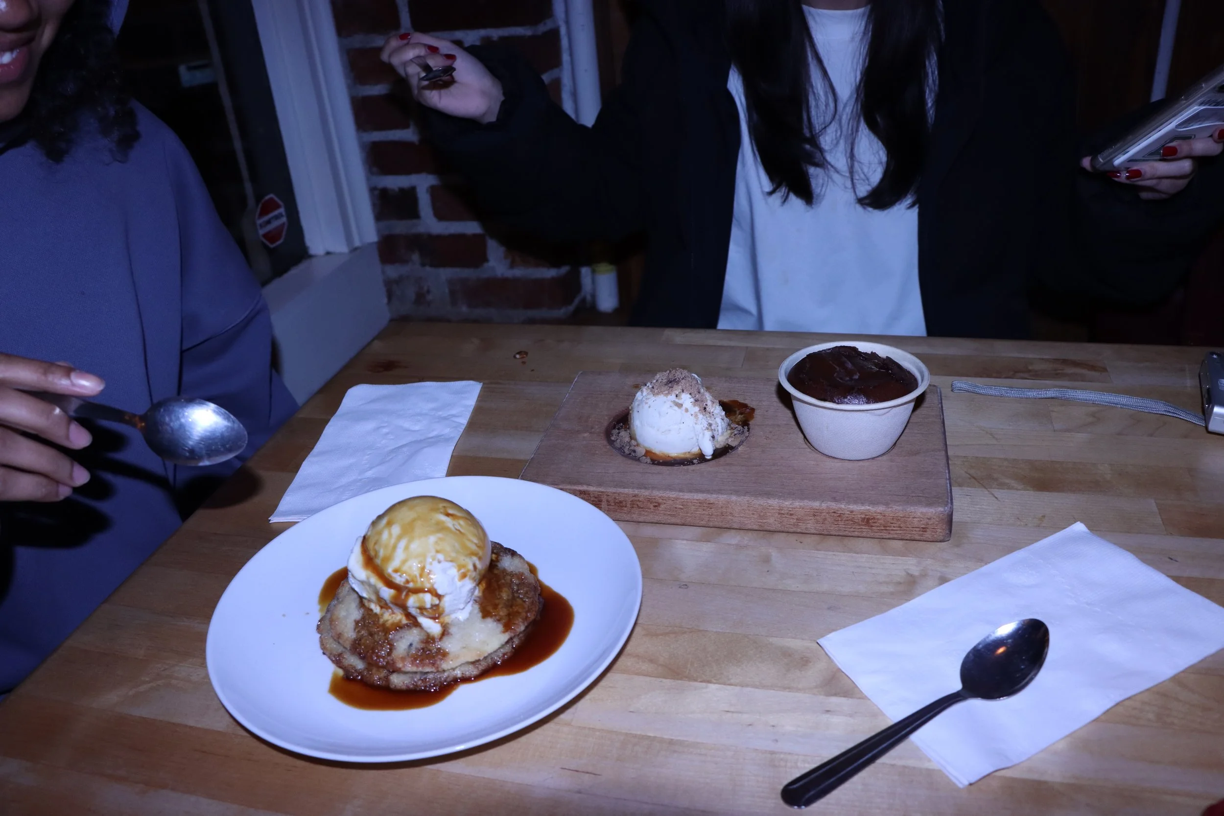 Desserts on a wooden table, including a plate with a dessert topped with a scoop of ice cream, a bowl of chocolate dessert, and two small desserts on a wooden board, with two people partially visible in the background.