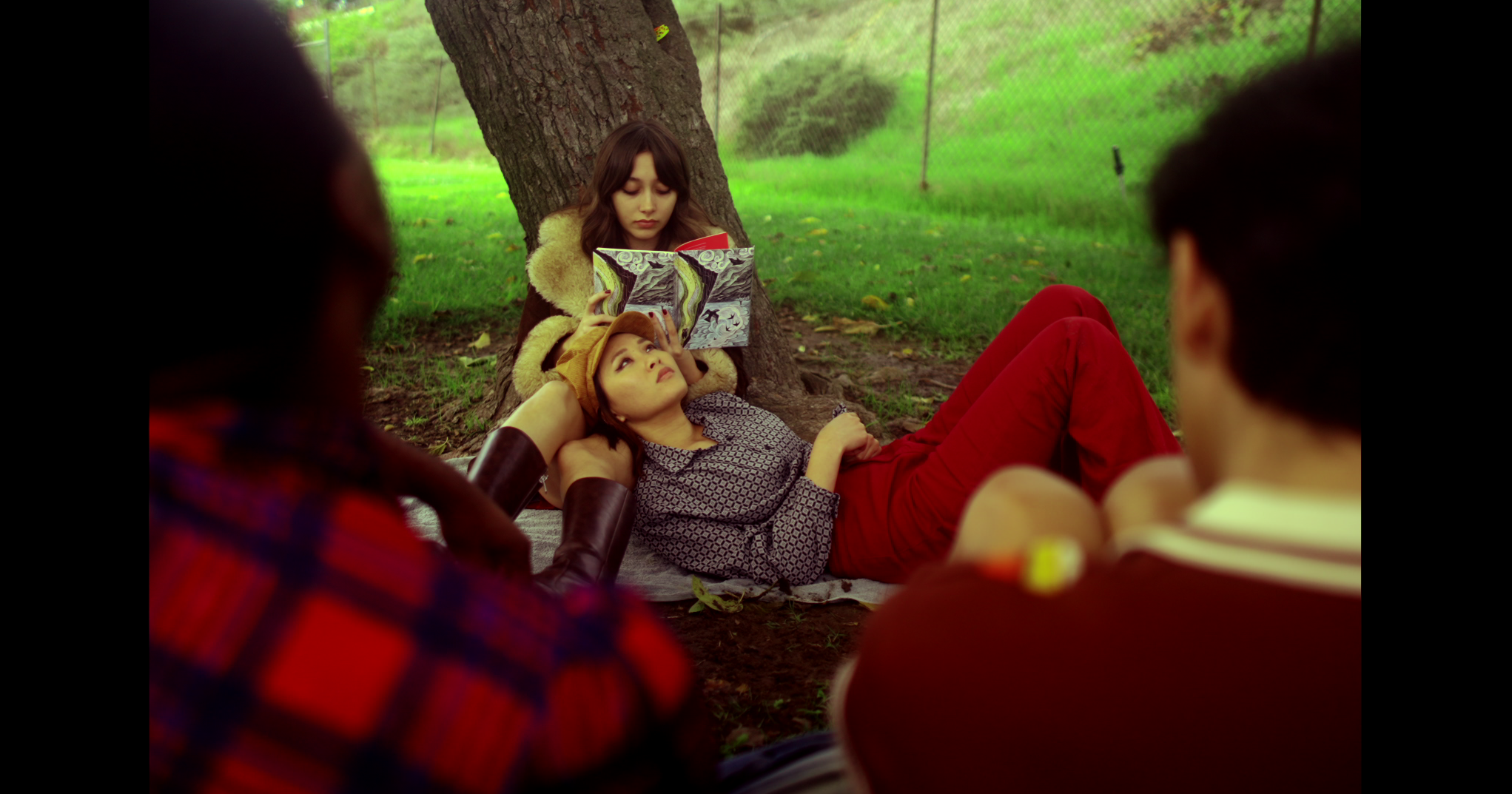 Young women sitting outdoors beneath a tree, one reading a book while another lies on the ground looking up, with two others blurred in the foreground, all in a grassy area with a wire fence in the background.