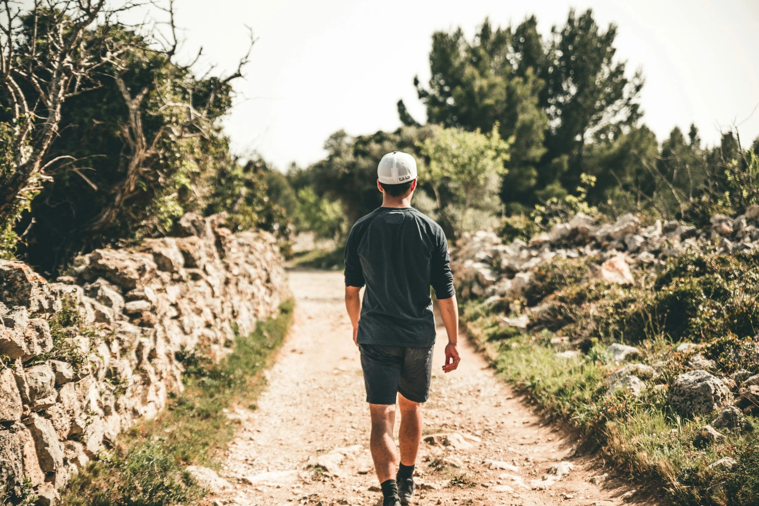 A person walking on a dirt path, surrounded by stone walls and green trees, during daytime.