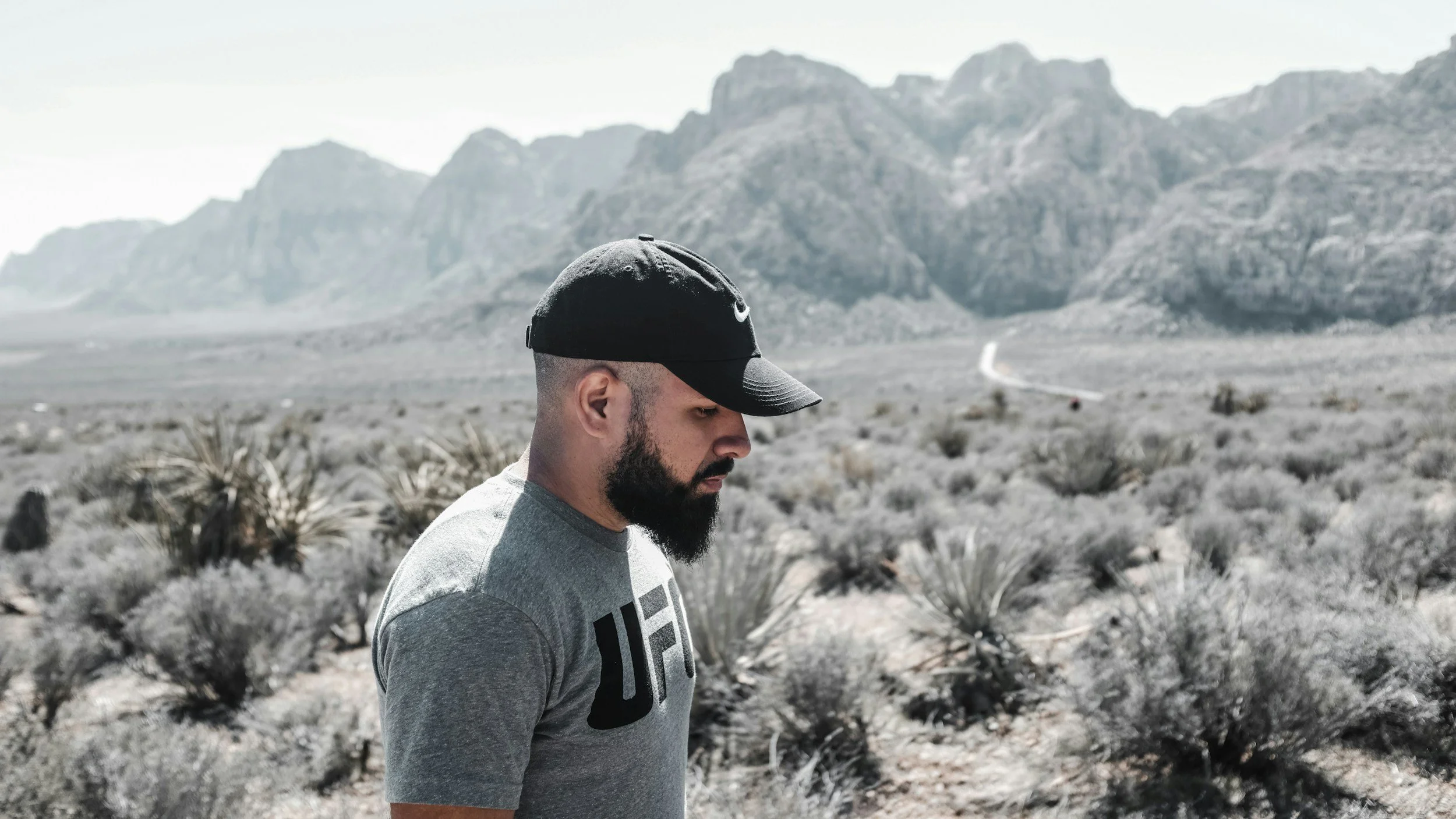 A man with a beard wearing a black Nike cap and gray t-shirt walking in a desert landscape with mountains in the background.