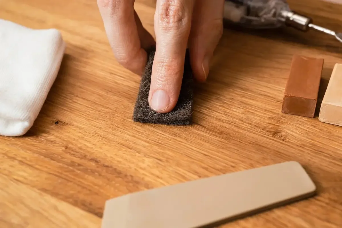 Close-up of someone using a black abrasive pad to polish or sand a wooden surface, with various tools and materials nearby.