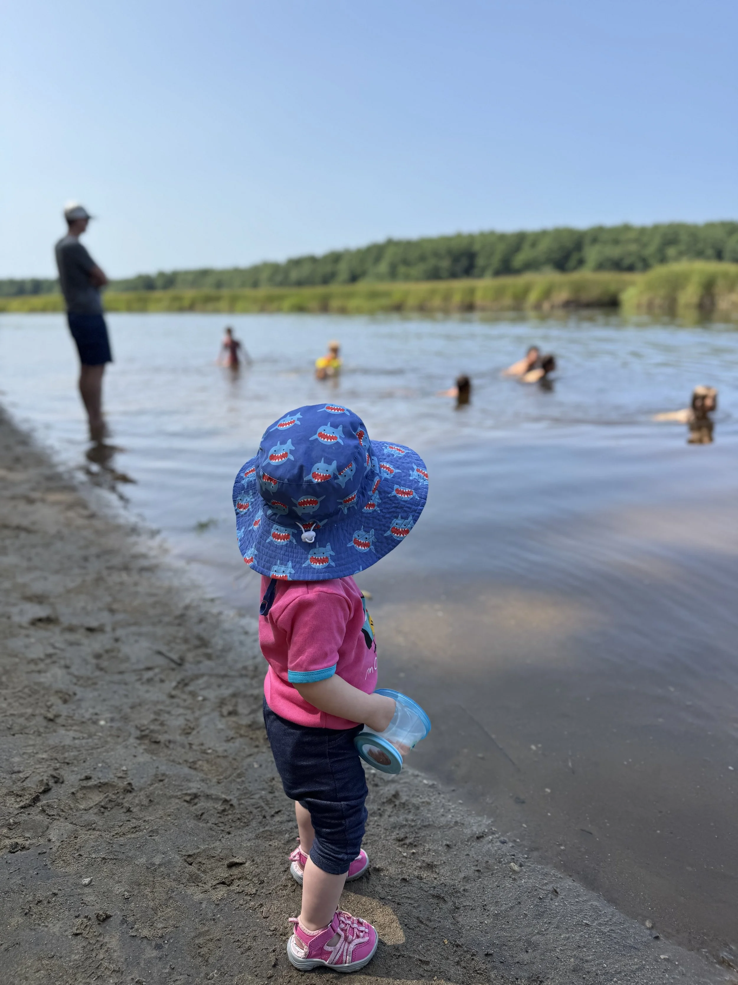 A toddler in a sunhat watches swimmers in a river as an adult supervises nearby.