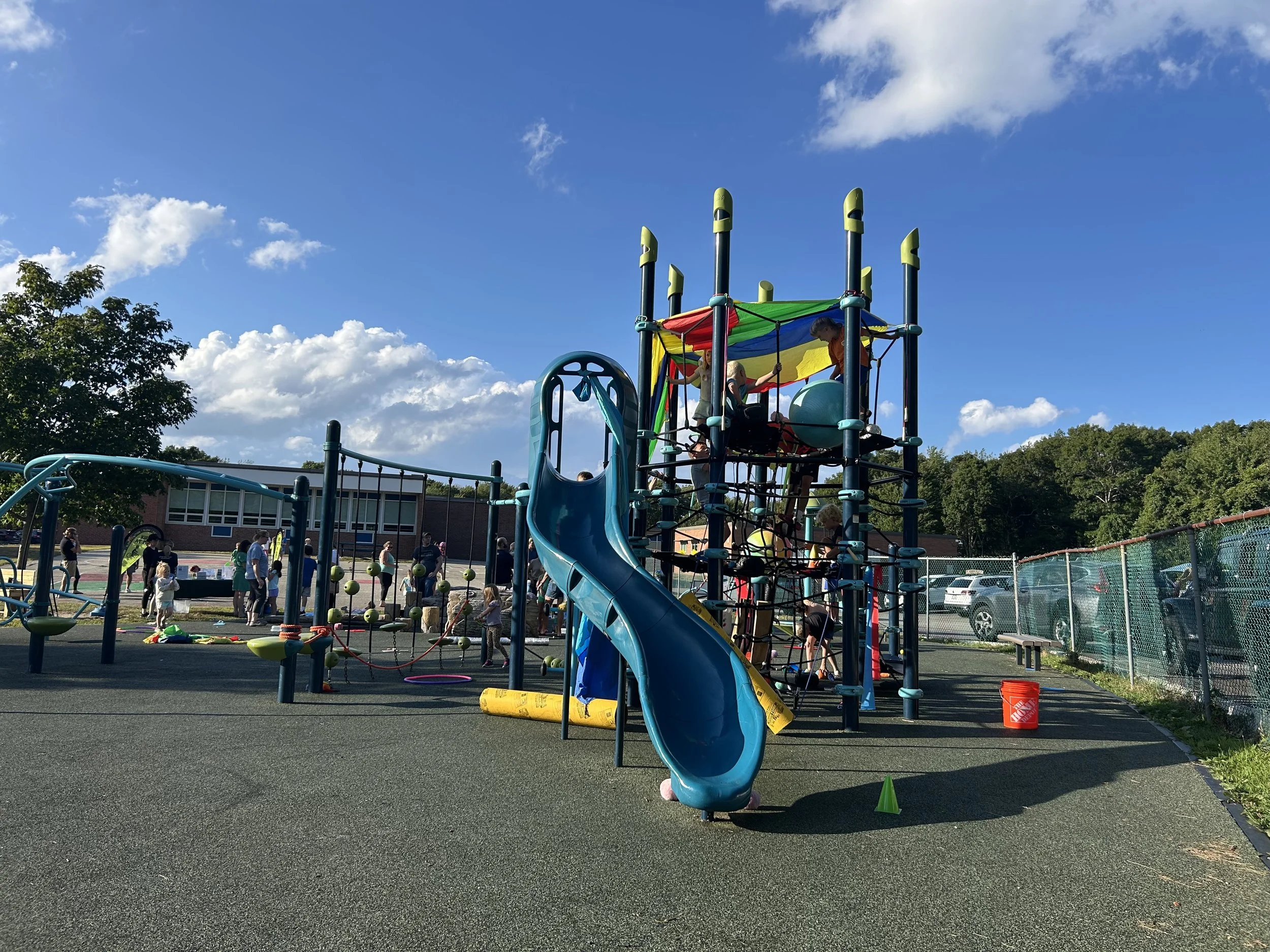 Under a bright blue sky, a play structure stands covered in large loose parts like tubes, cones, and play parachutes, with people in the distance.