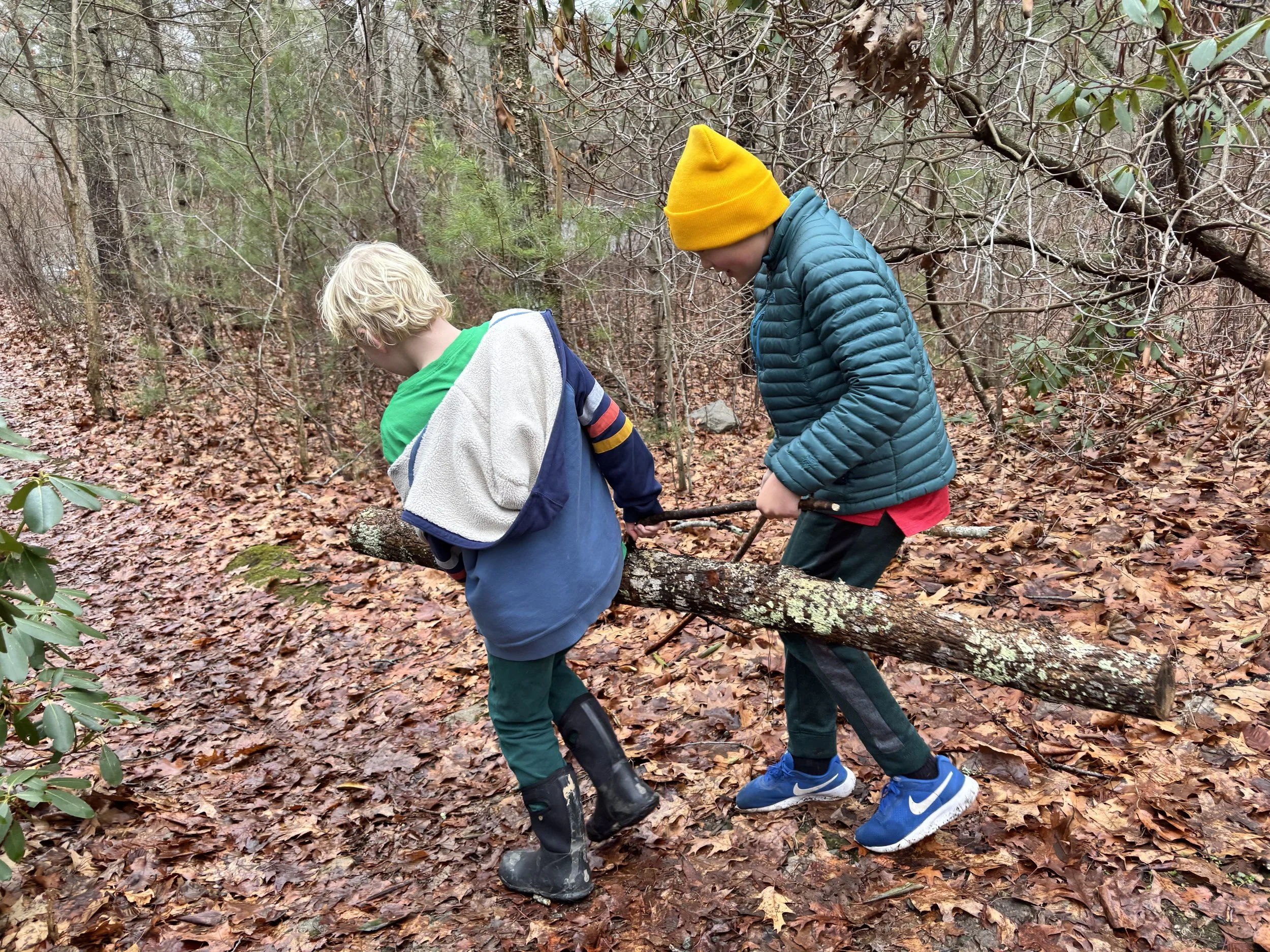 Two kids carry a heavy load together. They are outdoors lifting a log down a trail in the woods.