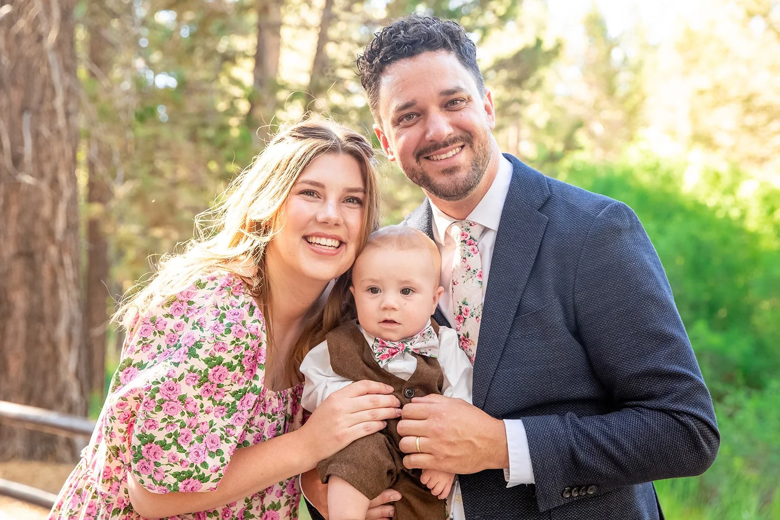 Betrayal Trauma and Postpartum Therapist. A smiling family of three, including a woman, a man, and a baby, outdoors in a wooded area with sunlight filtering through the trees.