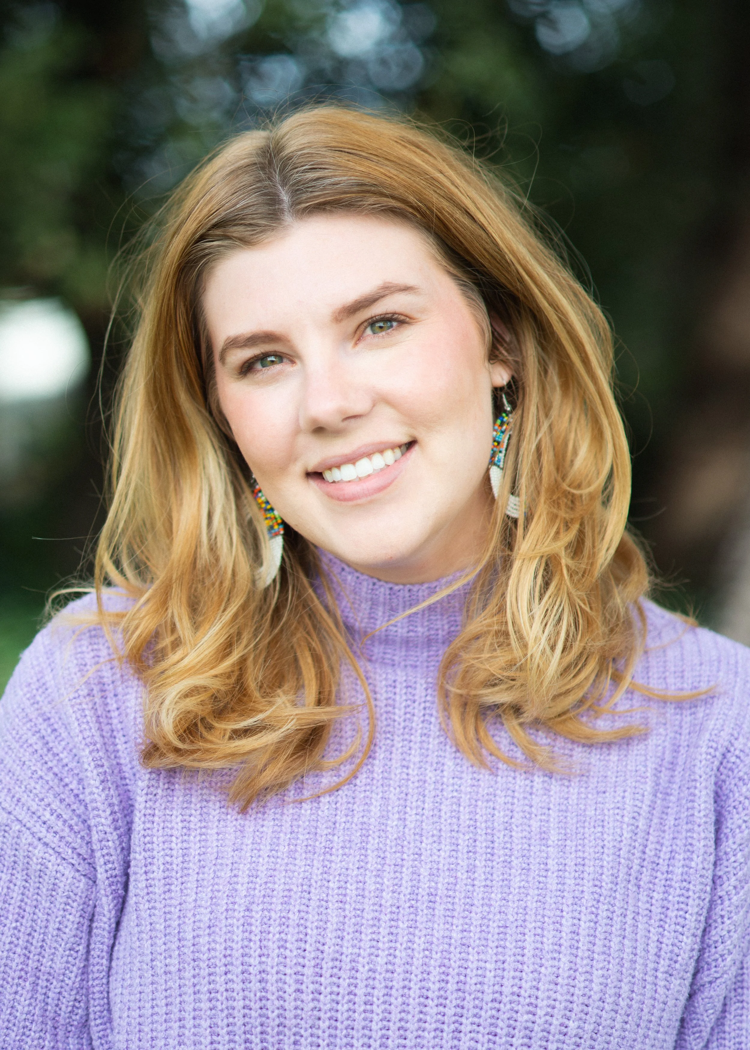 Betrayal Trauma and Couple's Therapist. Close-up of a young woman's face with long, wavy red hair, wearing a lavender sweater and beaded earrings, smiling outdoors with a blurred background of trees and greenery.