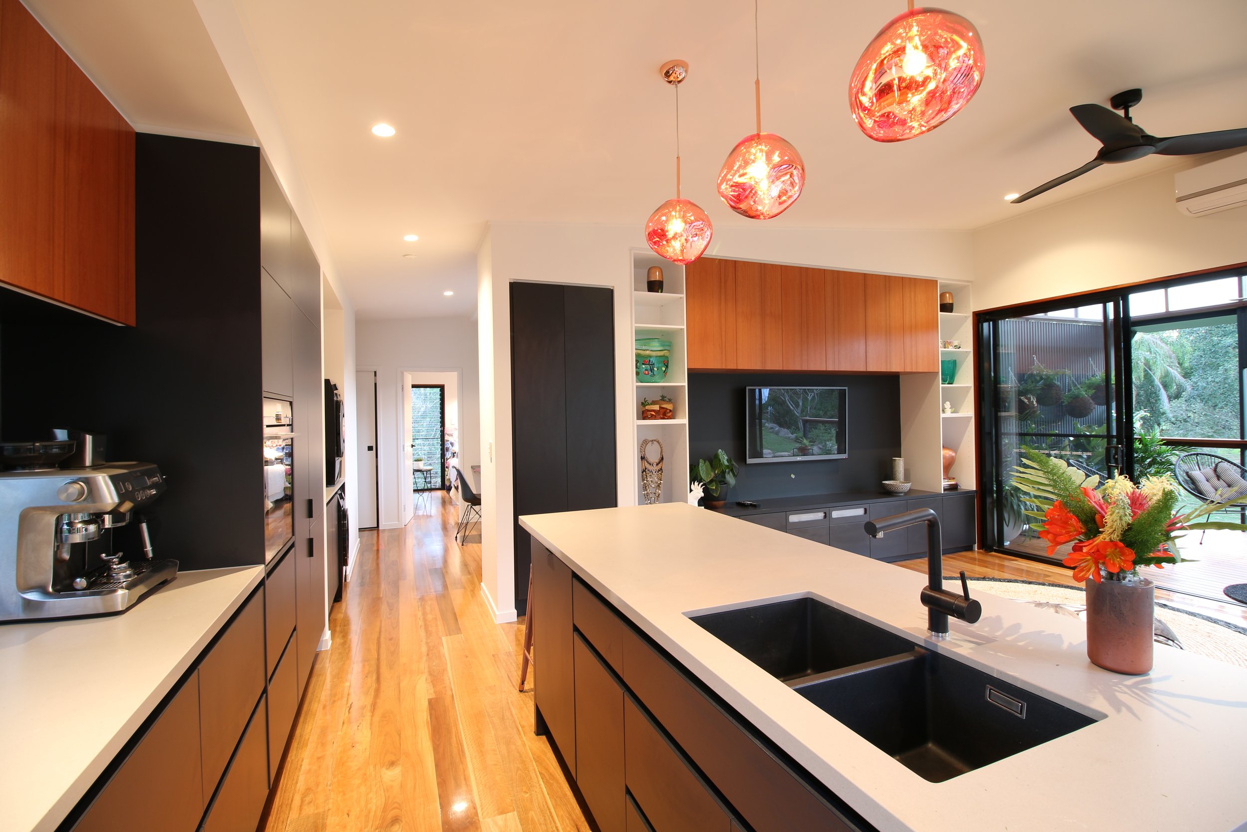 Modern kitchen with black cabinetry and white countertops, three red pendant lights, a black ceiling fan, sliding glass door opening to a backyard with outdoor seating and greenery.