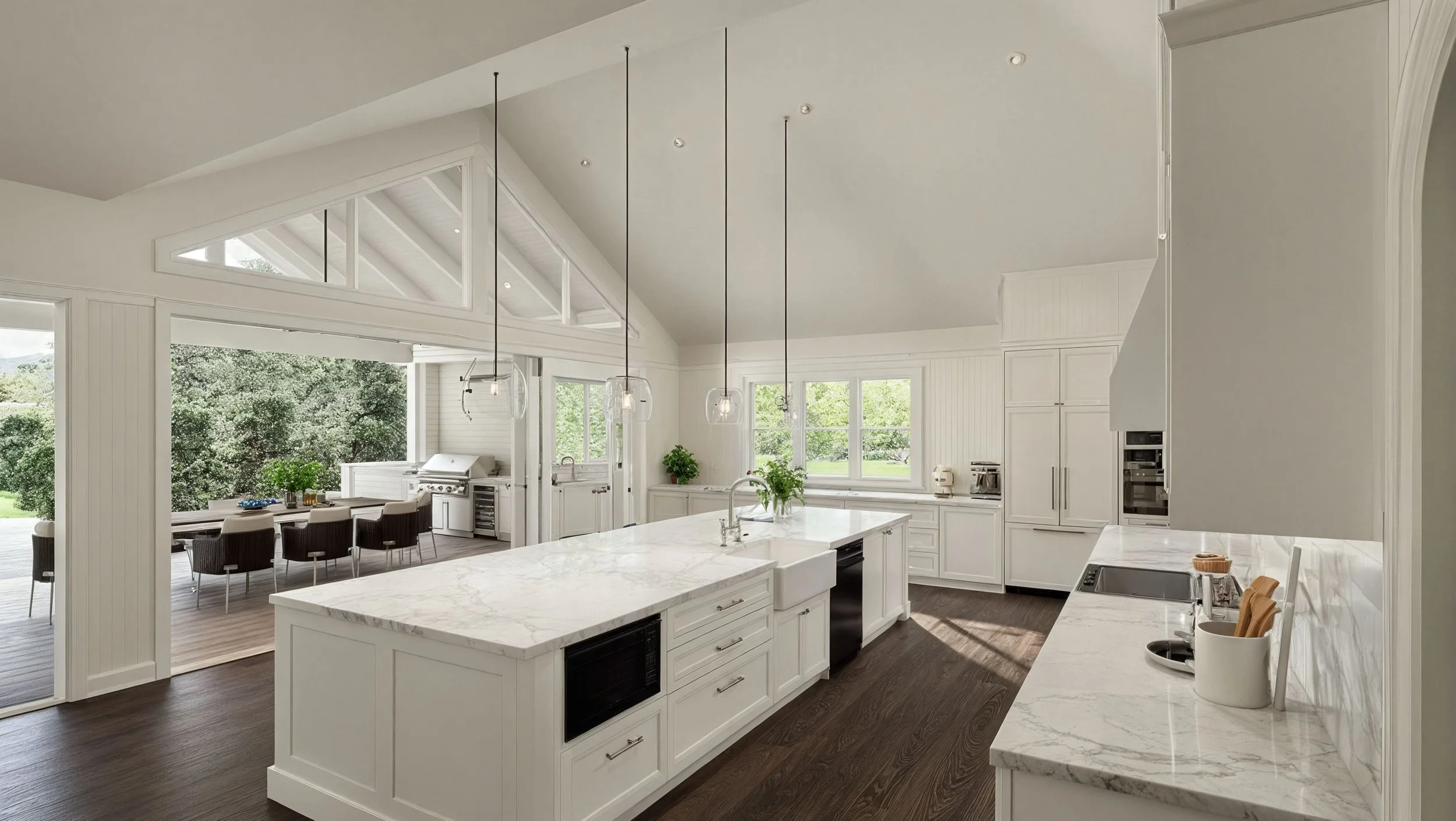 A bright, modern kitchen with white cabinets, marble countertops, and large windows overlooking greenery. There is a kitchen island with a sink, pendant lighting above, and a dining area with a rectangular table and chairs on a covered patio outside.