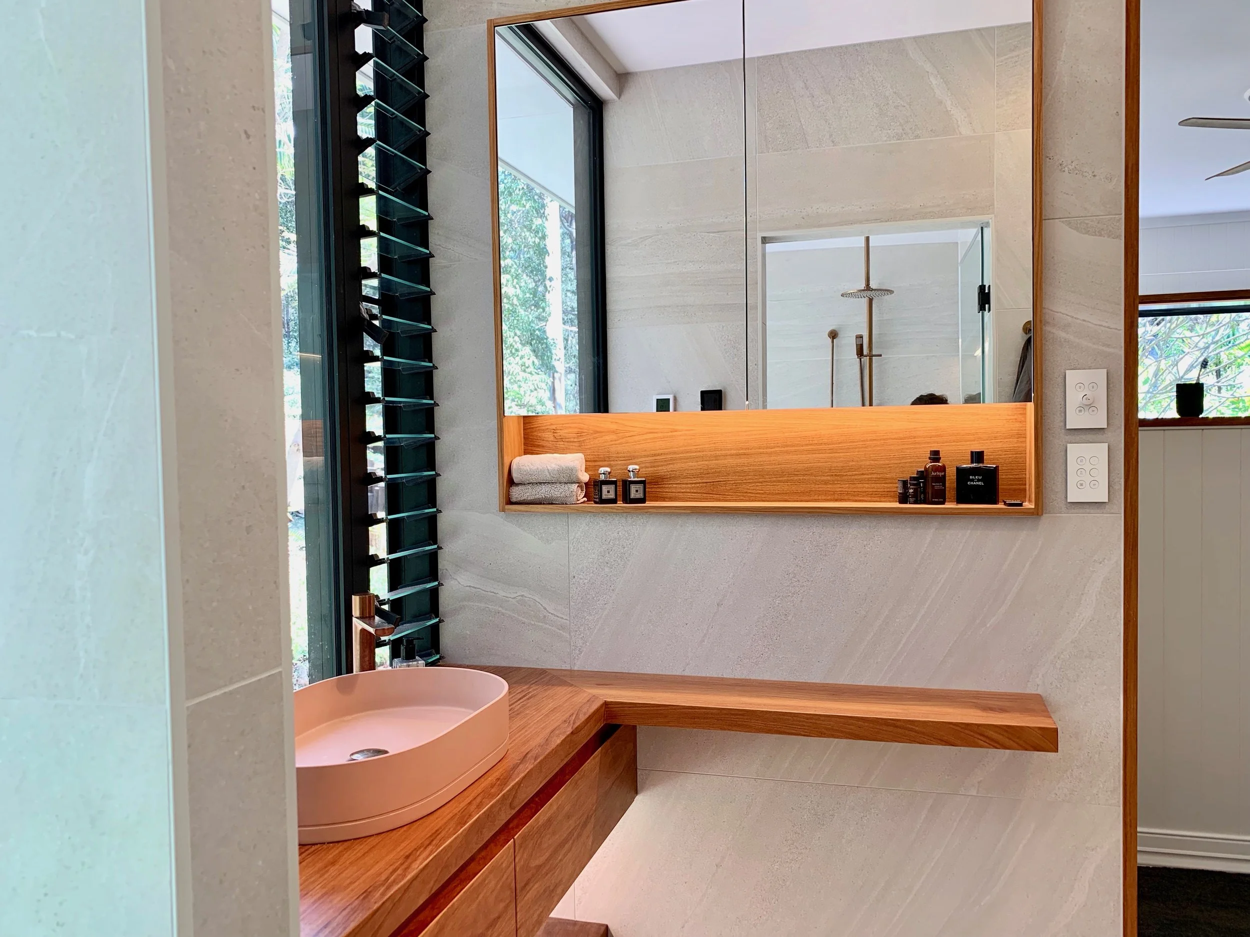 Modern bathroom with large mirror, beige stone walls, wooden countertop, pink vessel sink, and natural light from a window with trees outside.