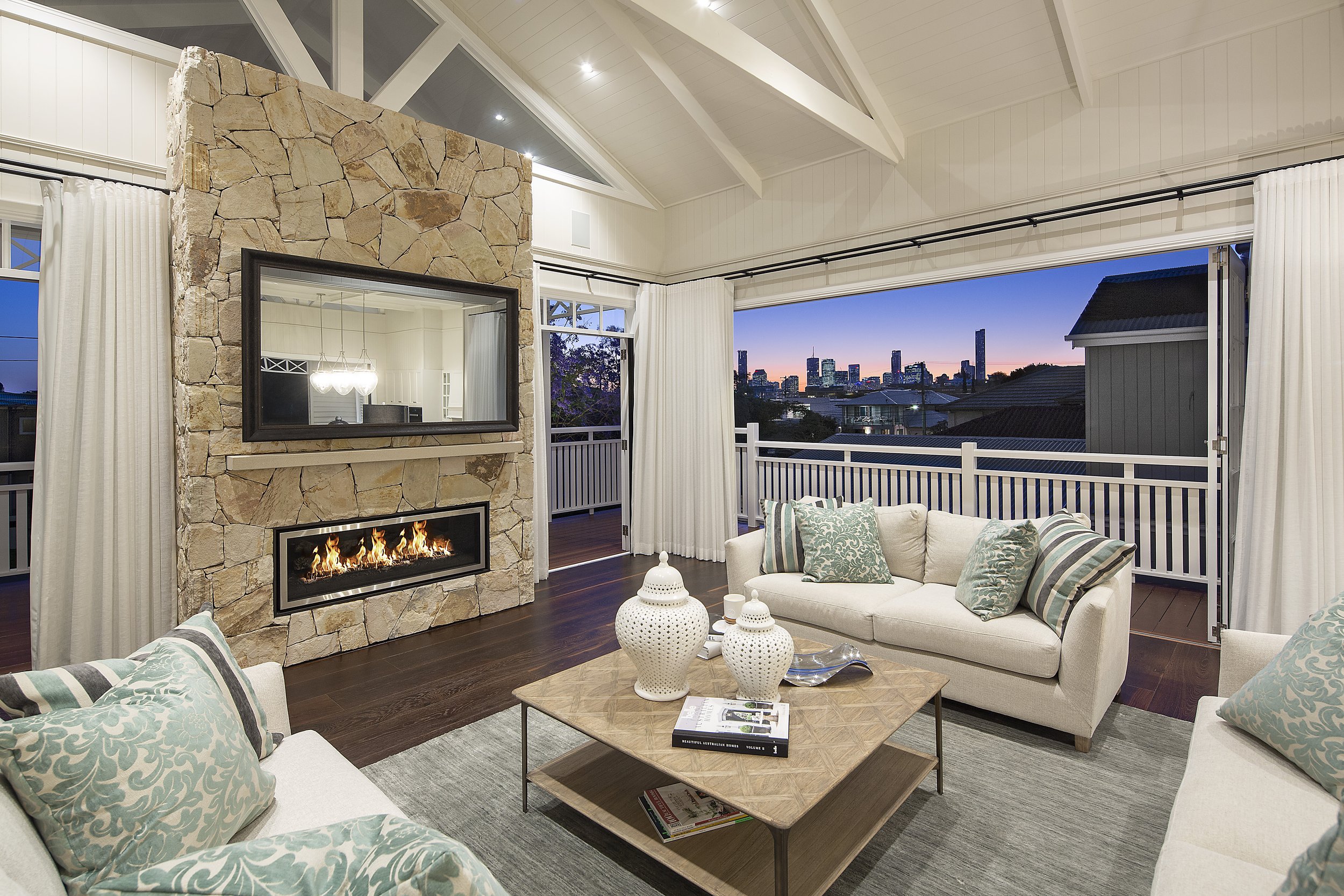 Living room with white sofas, teal patterned pillows, wooden coffee table with books and decorative items, a stone fireplace with a fire, large windows with white curtains, and a view of city buildings at sunset.