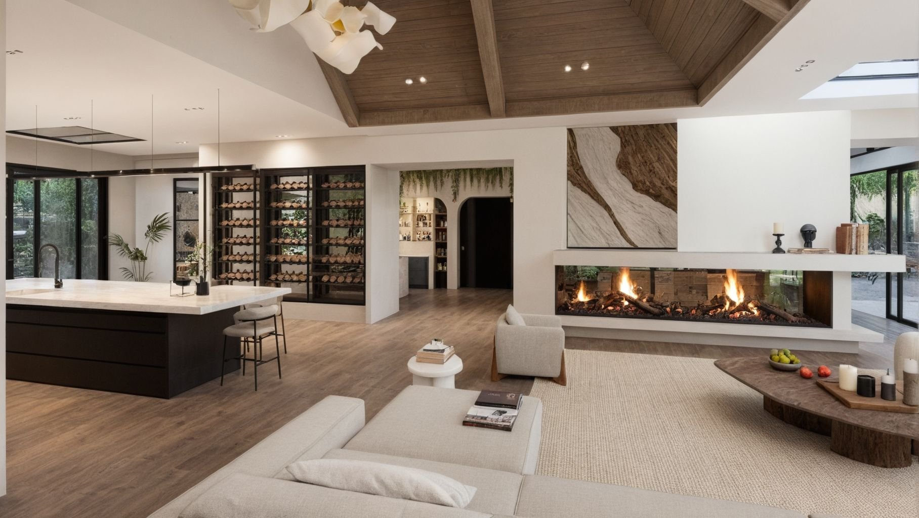 Modern living room with a double-sided fireplace, white walls, and large windows. The room features a beige sofa, a wooden coffee table with fruit, a built-in wine storage, and a kitchen island with bar stools. There are indoor plants, decorative can