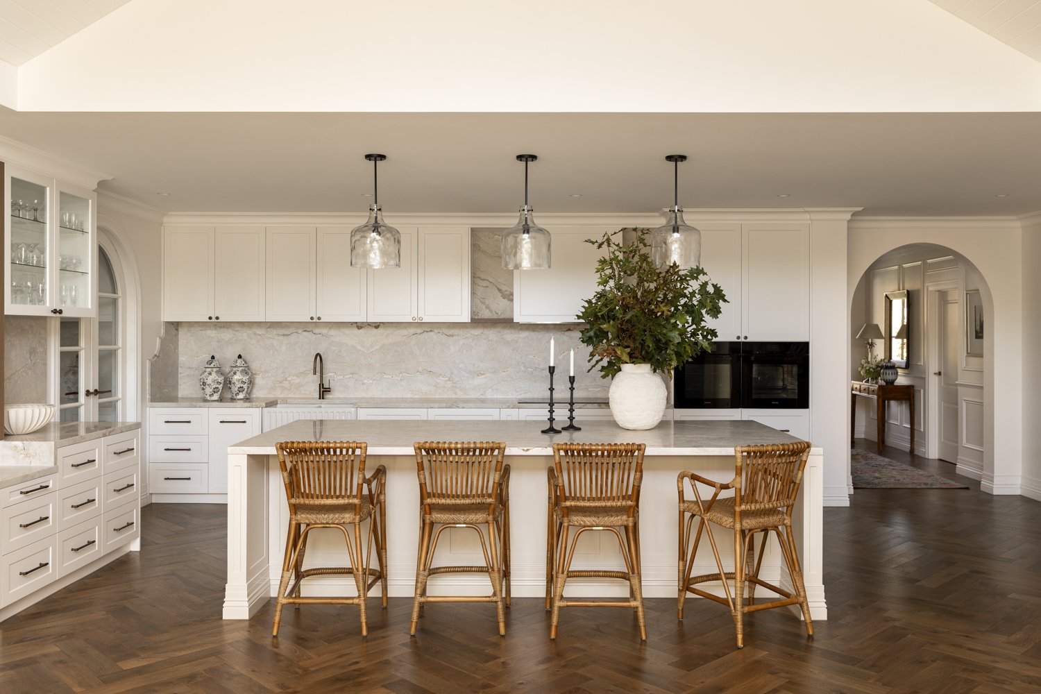 Modern kitchen with white cabinets, marble backsplash, central island with four wicker chairs, large vase with greenery, black candlesticks, and a built-in oven.