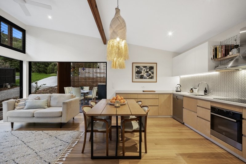 Open-concept living room and kitchen with modern decor, wooden flooring, a beige sofa, a dining table with fruit, a large window, and white cabinets.