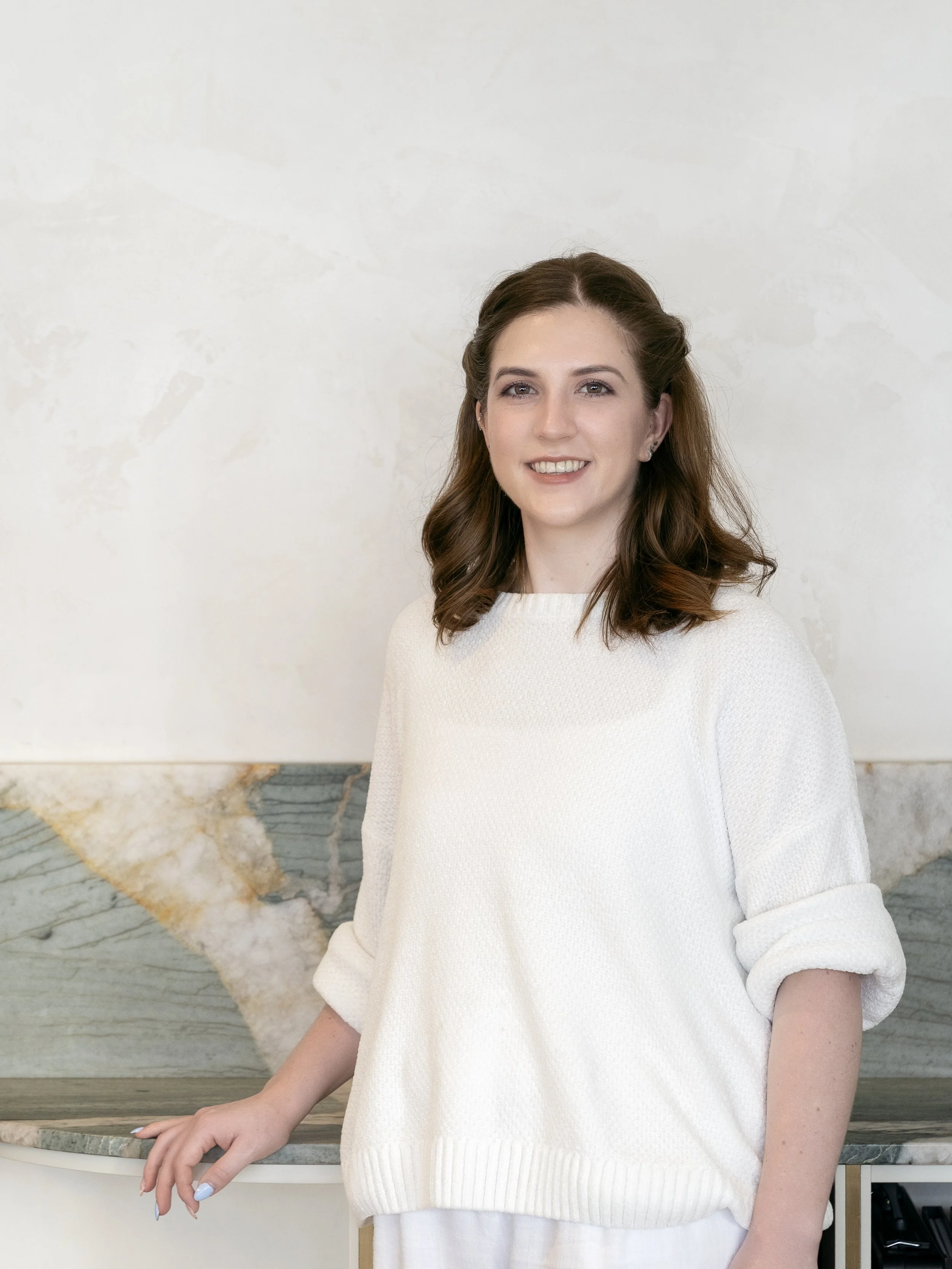 A woman with dark brown hair, partially pulled back, smiling and wearing a white sweater, standing indoors against a light-colored wall with a marble-like section and situated near a marble countertop.